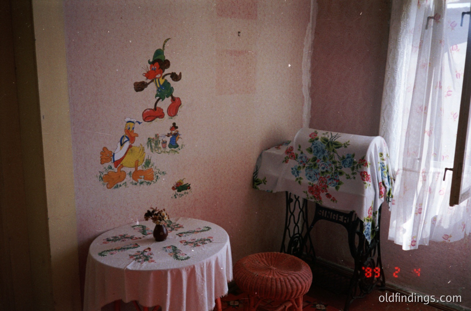 Vintage room featuring 1950s–60s Disney mural of Donald Duck, Mickey Mouse, and Goofy on faded wall. Round floral-patterned table with decorative pitcher and vintage sewing machine draped with floral cloth. Sunlight filters through lace curtains.