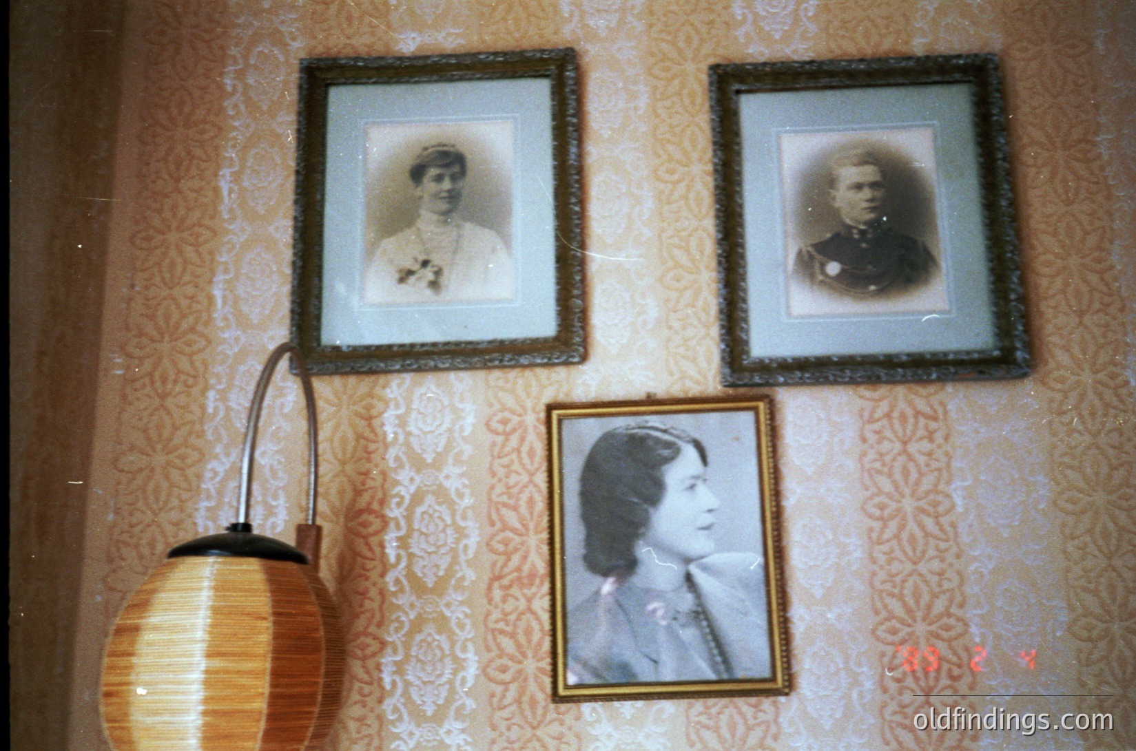 Vintage sepia-toned portraits in ornate frames on patterned wallpaper, likely early 20th century. Two framed images above show formal attire (women’s dress, military-style uniform), while a third portrait below depicts a woman in profile. A vintage hanging lamp with wooden slats adds warmth. Decor suggests a preserved family heirloom room.