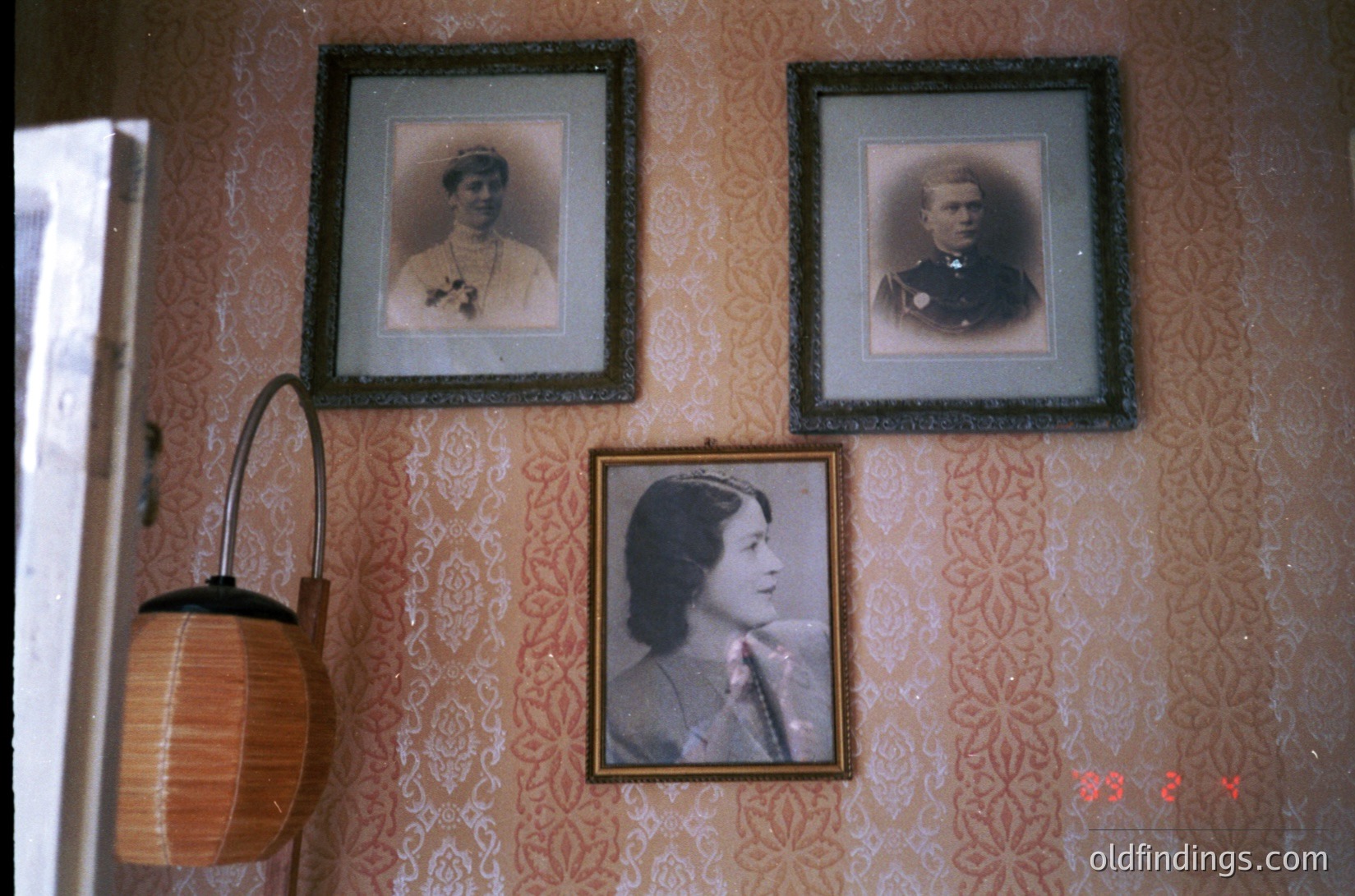 Vintage sepia-toned portrait gallery on ornate wallpapered wall, featuring three framed photos in ornate frames. Top left: woman in early 20th-century dress with floral pattern; top right: man in military uniform with insignia; bottom center: woman in high-necked blouse. Wicker wall lamp with curved handle at left. Likely late 19th to early 20th century European interior.