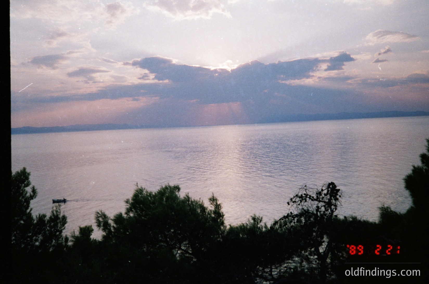 Sunset over a vast body of water with distant landmass, framed by greenery. Soft lighting casts reflections on calm waters. Likely a coastal or lakeside scene, possibly due to hue and horizon. Vintage photo stamp suggests or early .