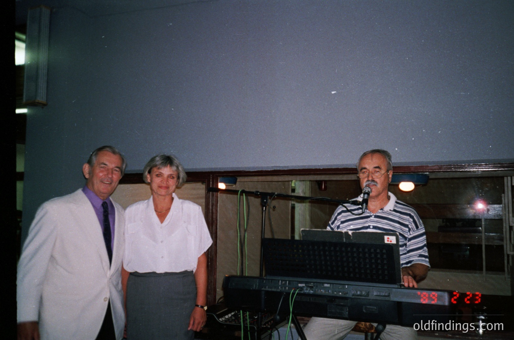 Indoor event featuring three adults posing near a keyboard with digital clock displaying "23:23." Man on left wears a white lab coat with purple tie; woman in center wears a light blouse; man on right wears a striped shirt and holds a microphone. Dim lighting, likely a 1990s-2000s venue.