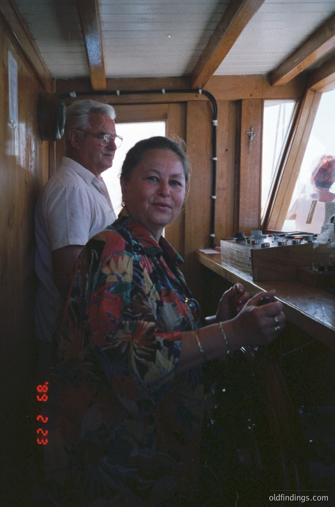 Indoor scene featuring two adults in a rustic wooden cabin. The woman, wearing a floral-patterned blouse, stands at a wooden counter with a vintage radio or clock (marked "33 23") beside her. The man in a light-colored shirt leans against the counter. Natural light streams through large windows, illuminating the wooden beams and interior. Likely mid-20th century, rural or coastal setting.