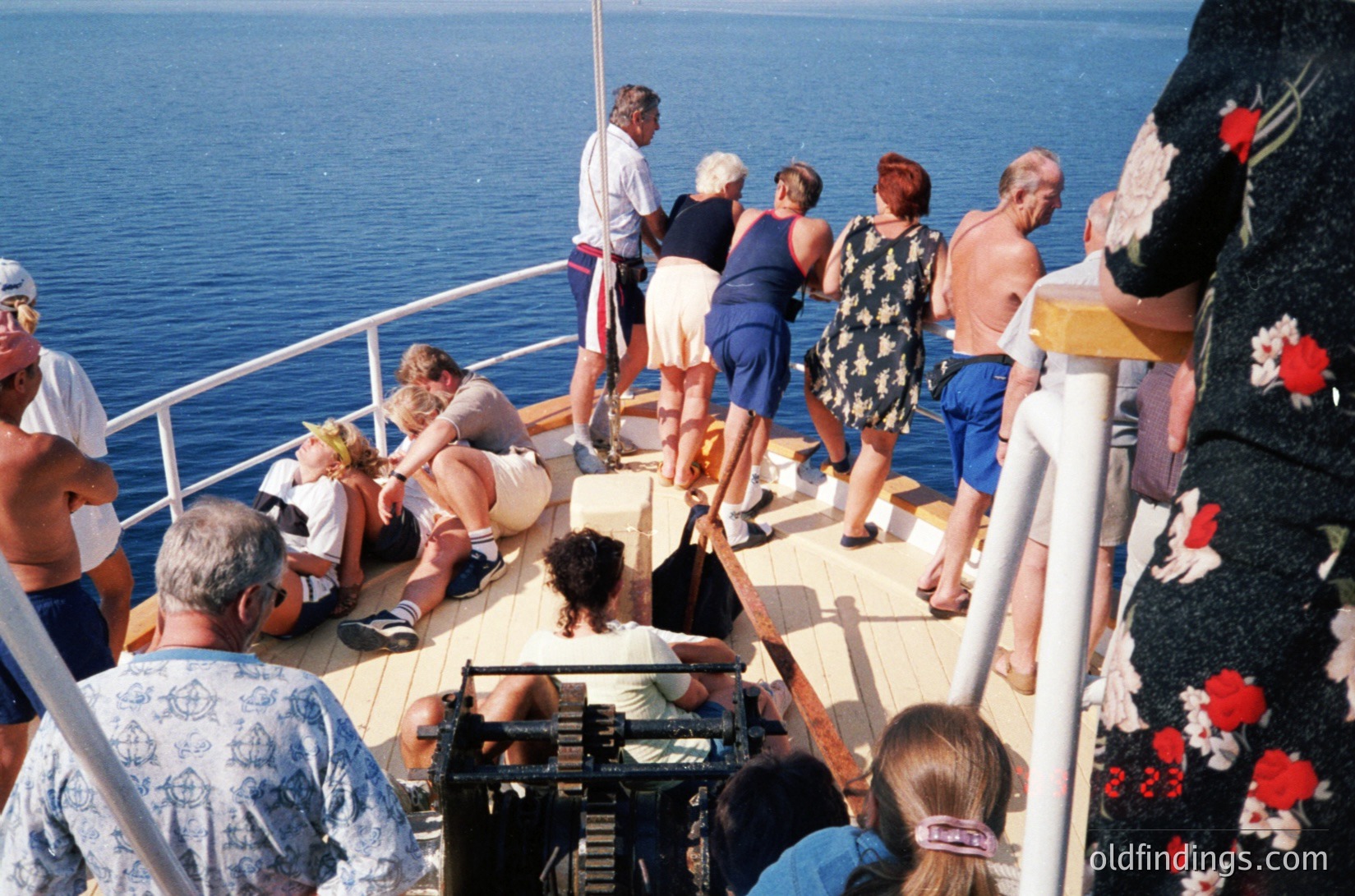 Mid-1970s cruise ship deck scene: adults in retro swimwear (one-man suit, floral dresses) gather on sun deck by railing, some leaning over edge. Bright blue sea and clear skies in background. Decorative embroidered fabric (red floral pattern) visible at right edge.