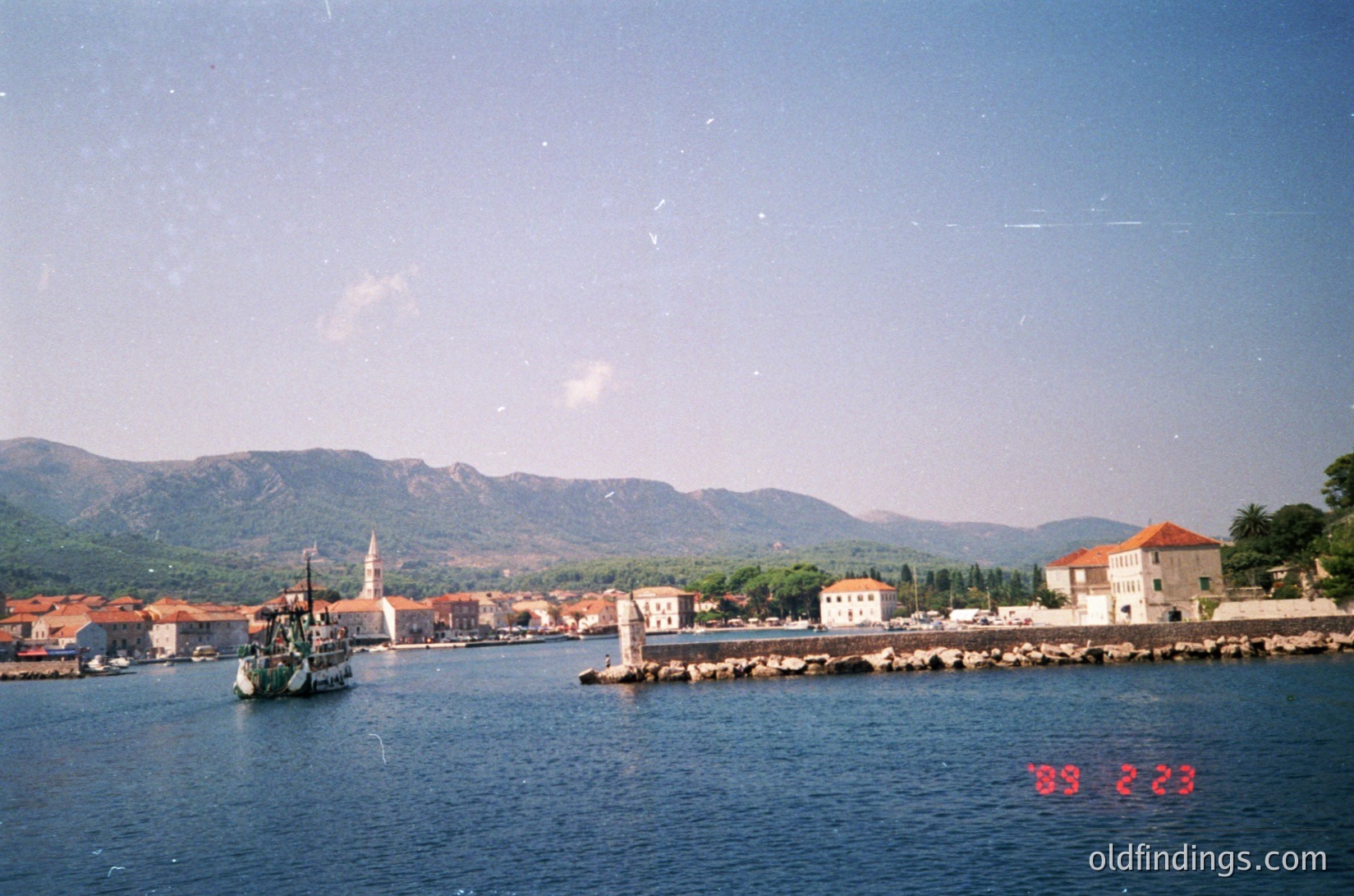 Vintage coastal town with Mediterranean architecture—whitewashed buildings, terracotta roofs, and a prominent church steeple. A traditional wooden sailboat docked near a stone pier, framed by calm waters and mountainous backdrop. Likely Adriatic Sea region, mid-20th century.