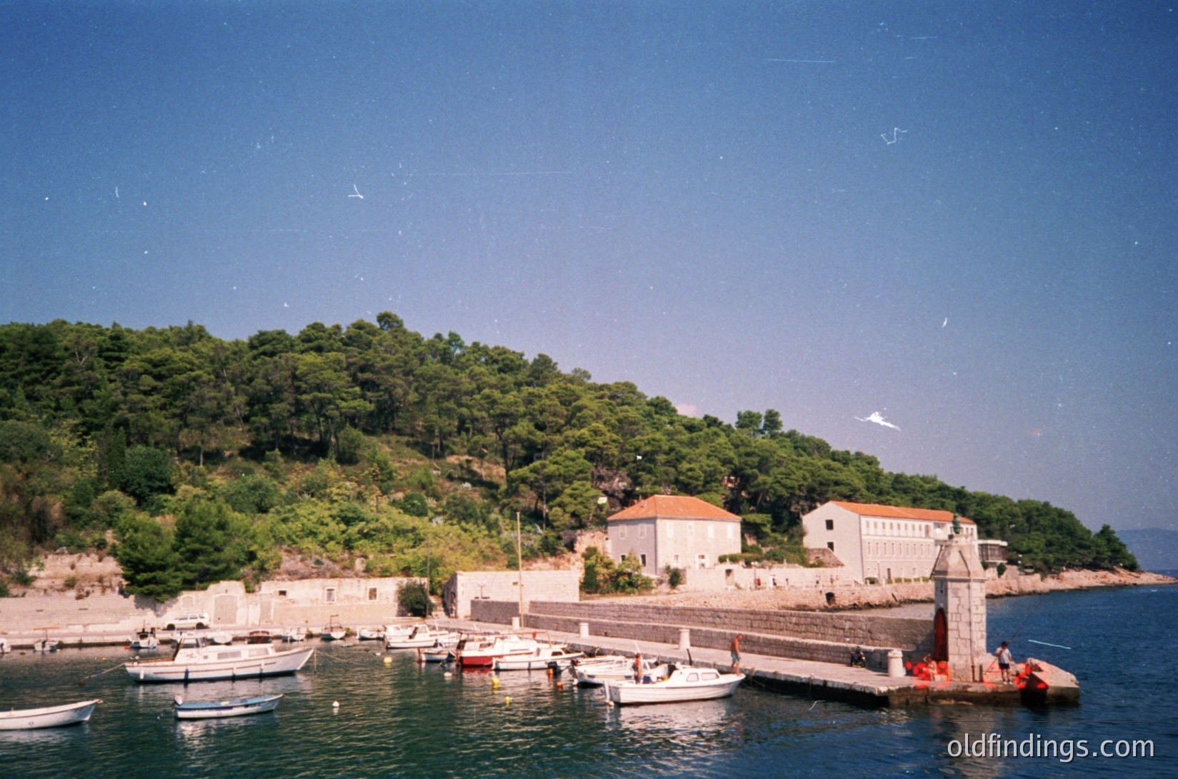 Coastal village scene with stone buildings perched on a hillside, overlooking a small harbor. White boats docked at a stone pier extend into deep blue waters. Lush green forest covers the hill, contrasting with the clear blue sky. Likely Mediterranean, possibly Adriatic region.
