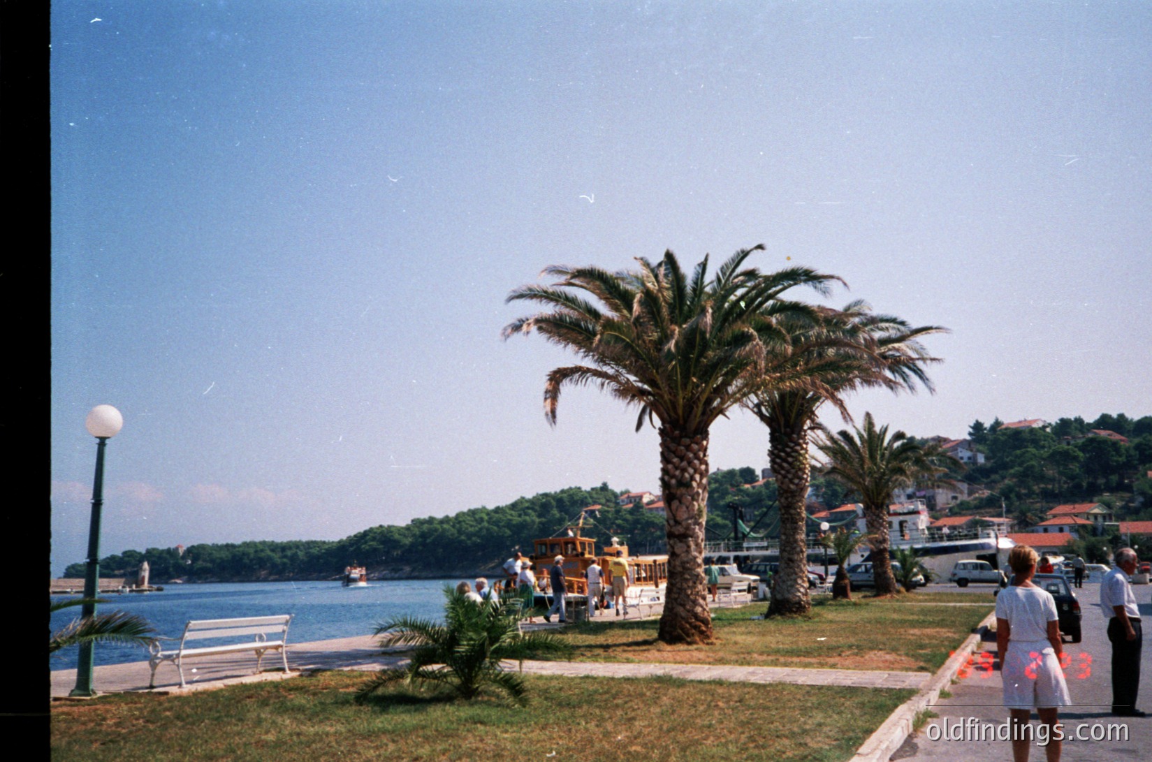 Vintage seaside promenade with palm-lined walkway, likely Mediterranean. Clear blue water reflects sunlight; boats docked near shore. Mid-20th century architecture in background. Empty benches and a lone figure in white suggest quiet, possibly early tourism era.