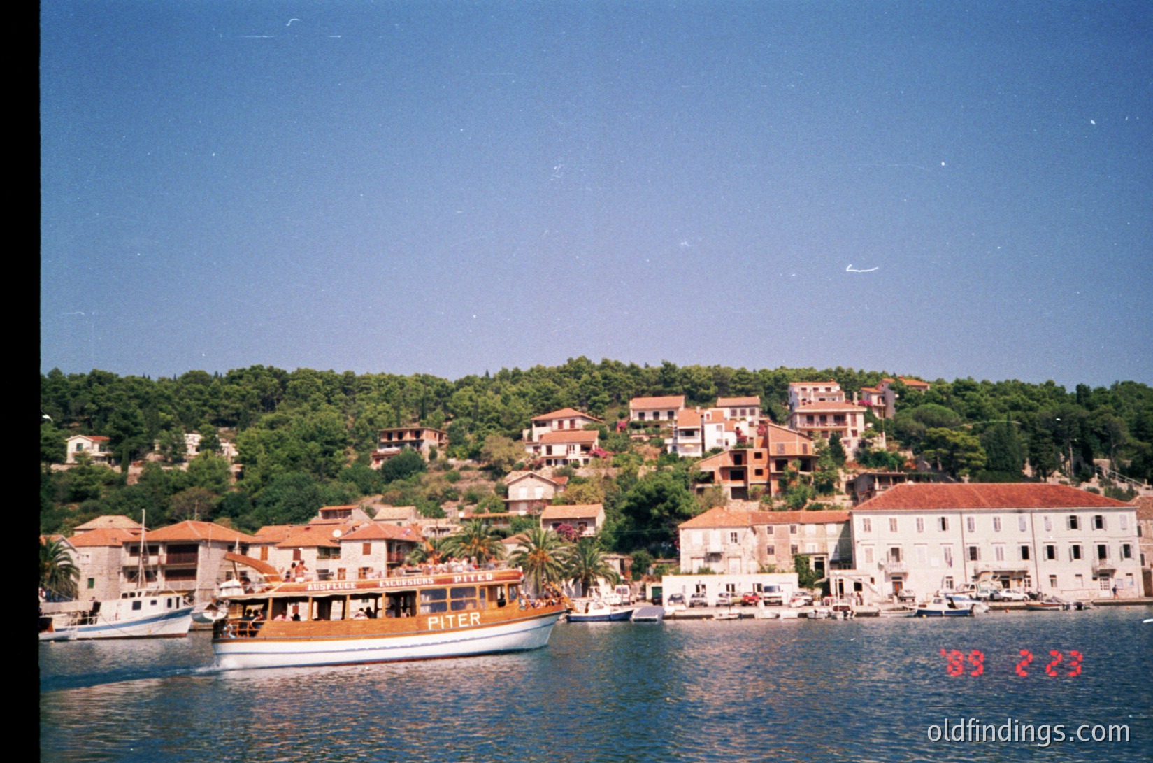 Mediterranean coastal village with clustered stone buildings and terracotta roofs. A vintage ferry labeled "Piter" docked near a small harbor, surrounded by calm blue waters. Lush greenery and hills frame the scene, suggesting a Mediterranean climate. Likely due to film grain and ferry design.