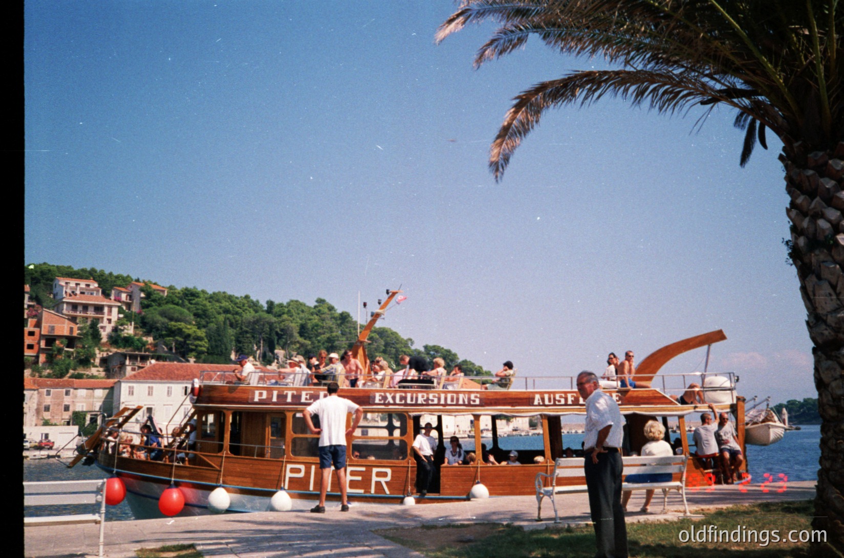 Vintage wooden excursion boat named **"Piter"** docked at a seaside port, labeled "Excursions" in bold letters. Mid-20th century Mediterranean coastal scene with stone buildings and lush greenery. Clear skies and palm trees frame the bustling dockside activity.