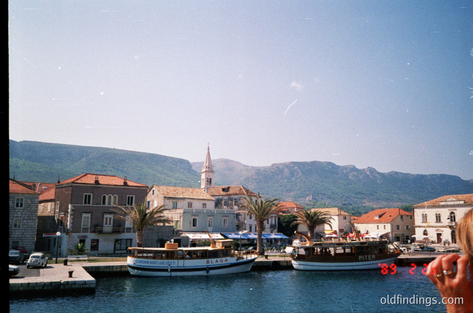 Mediterranean coastal town with stone buildings, palm trees, and a church steeple. Two boats docked near the waterfront, one labeled "Piter." Mountain backdrop suggests a sheltered harbor. Likely Adriatic coast, 1980s–1990s.
