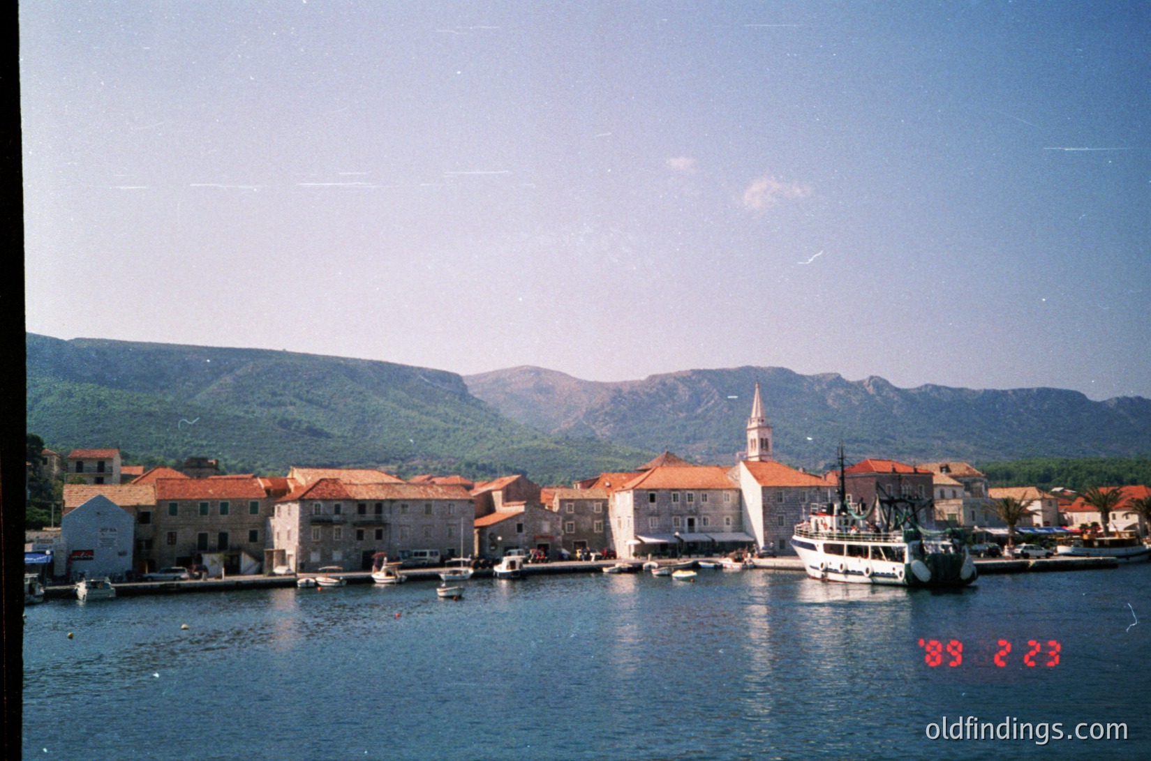 Mediterranean coastal village with stone buildings, terracotta roofs, and a prominent church steeple. Small boats docked along the waterfront, framed by rugged hills. Likely