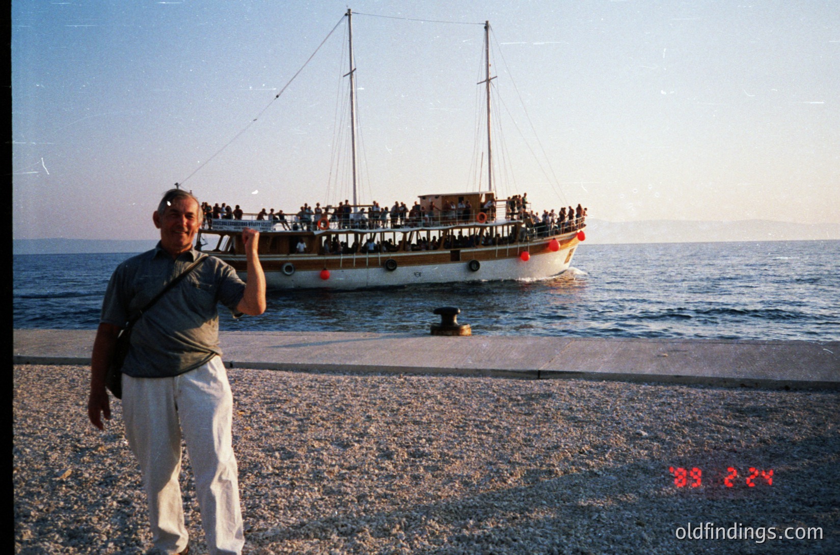 Vintage seaside scene featuring a man in retro attire (striped shirt, light pants) posing on a pebble shore. Behind him, a wooden passenger ferry packed with people cruises near the coast. The ship’s red life buoys and classic design suggest mid-20th century maritime transport. Likely Mediterranean or Black Sea region.