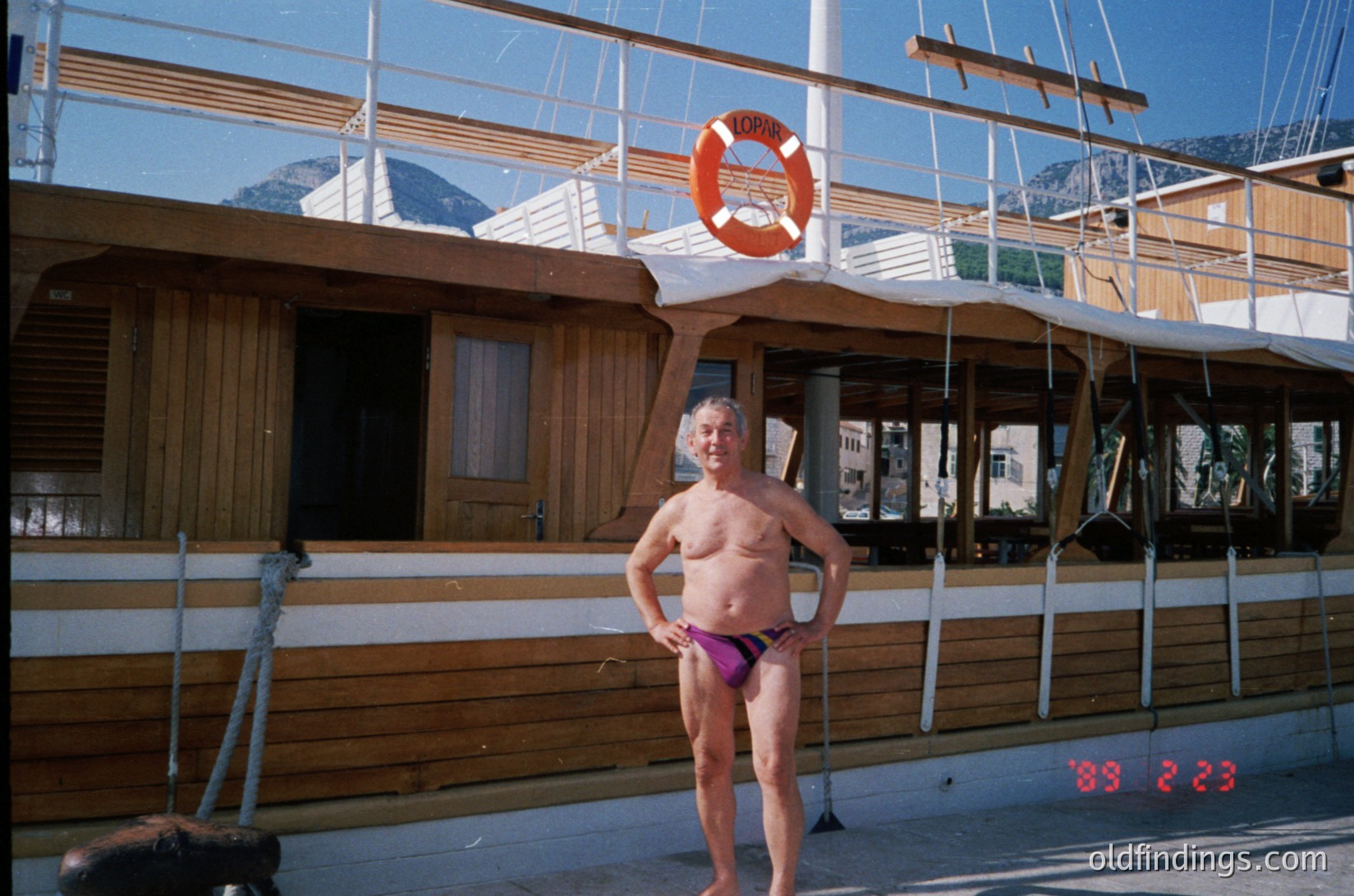 Vintage seaside scene featuring a shirtless man in 1970s-style swim trunks posing beside a wooden docked boat. The vessel displays a prominent orange lifebuoy with "LOFOTEN" inscribed. Mountainous backdrop suggests a coastal Alpine or Nordic region. Dock number "88 223" visible in red.