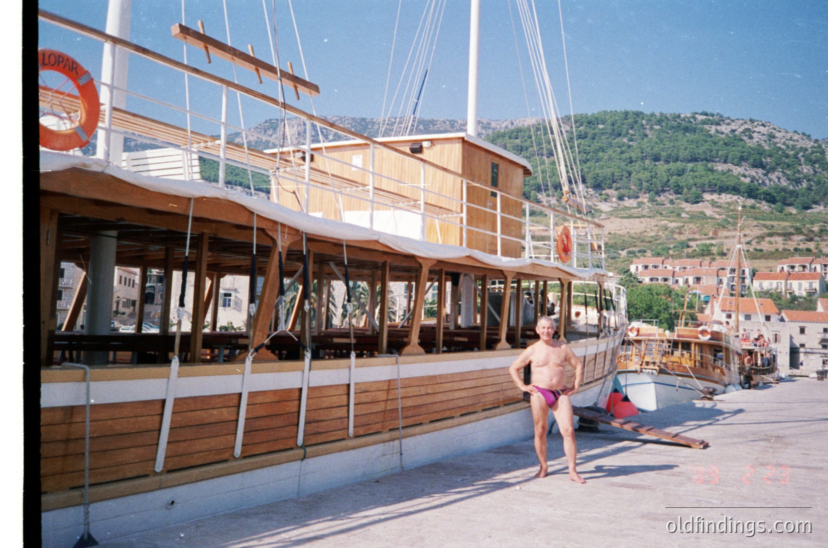 Classic wooden sailing yacht docked at a Mediterranean port, featuring a high deck with railings and lifebuoys. A shirtless man in pink swim trunks stands near the stern, with mountainous terrain and coastal village in background. Likely Mediterranean seaside scene.