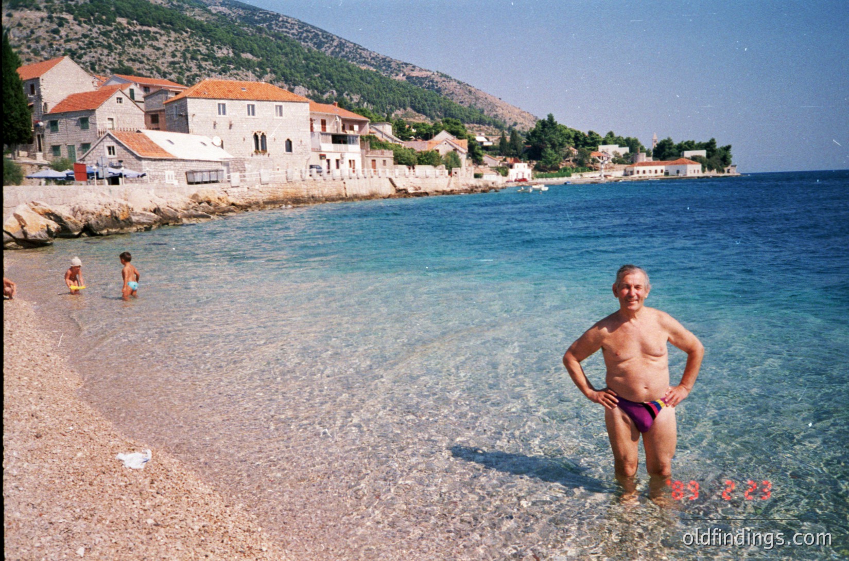 Vintage seaside scene featuring a man in red swim trunks standing in shallow turquoise waters, with Mediterranean-style stone buildings and greenery-lined cliffs in background. Likely coastal Croatia or Dalmatia, 1970s-1980s.
