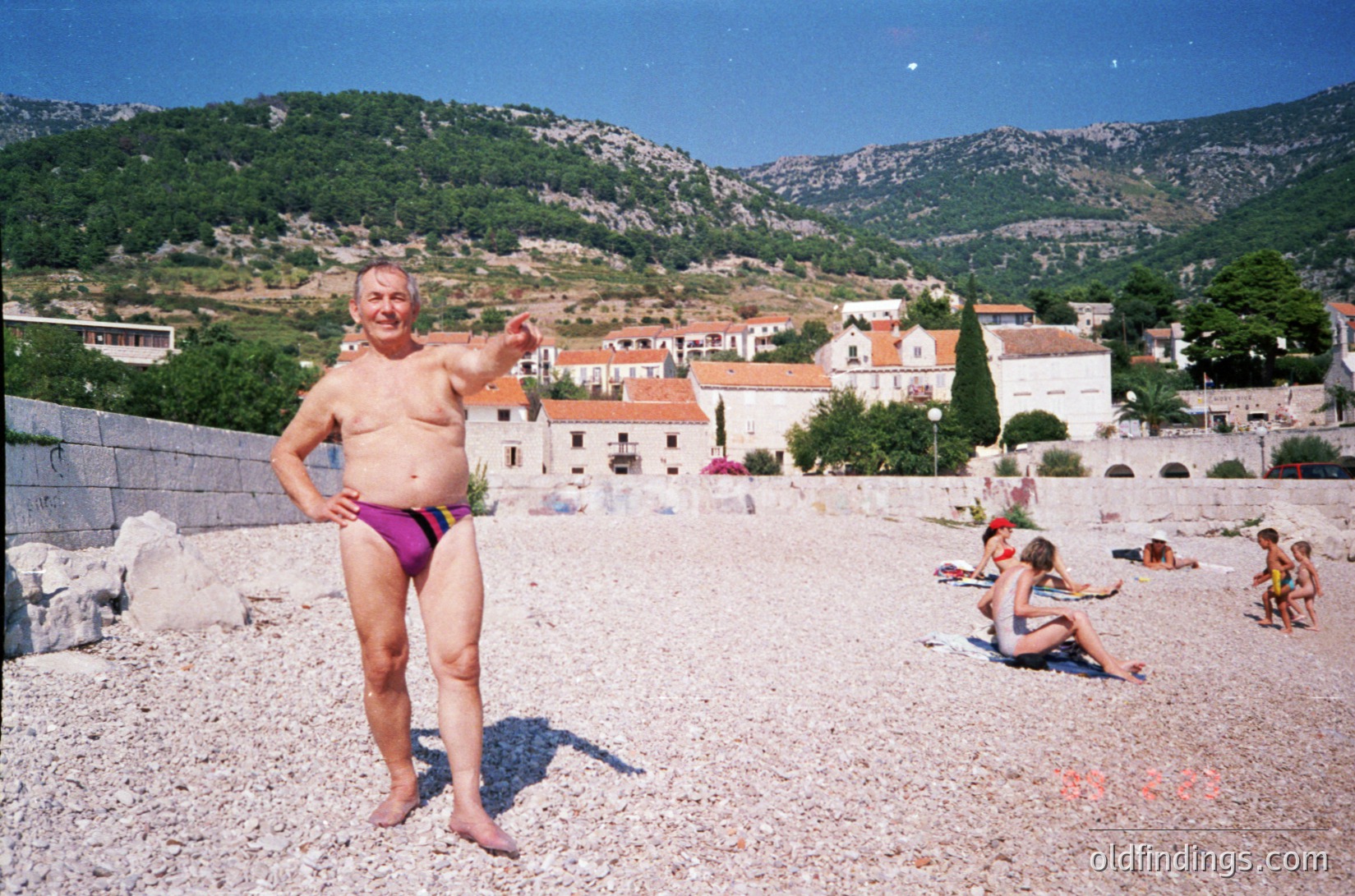 Shallow gravel beach with Mediterranean coastal village in background. Elderly man in purple swim trunks poses confidently, gesturing. Midday sun casts clear shadows. Likely seaside