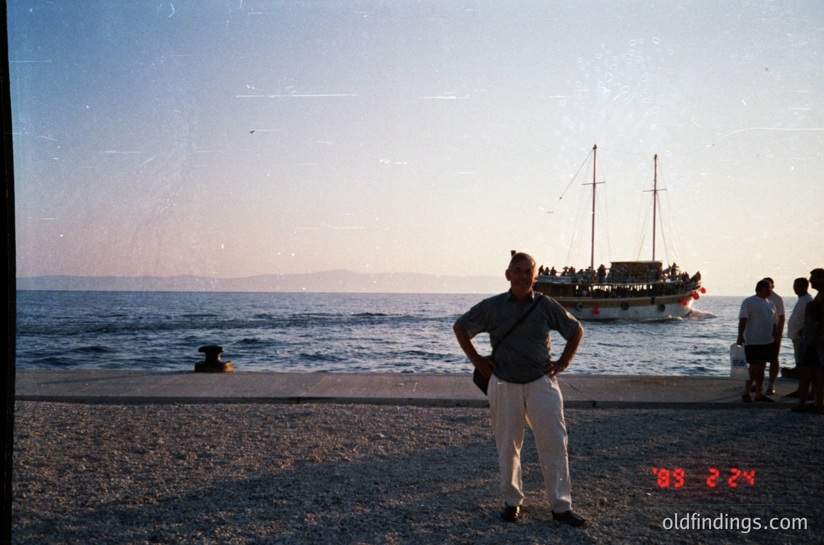 Vintage seaside scene with man in light-colored trousers and dark polo standing on pebble shore. Wooden pier extends into calm waters with a traditional wooden boat anchored nearby. Faint date stamp "1992" visible. Coastal landscape includes distant landmass under golden-hour light.