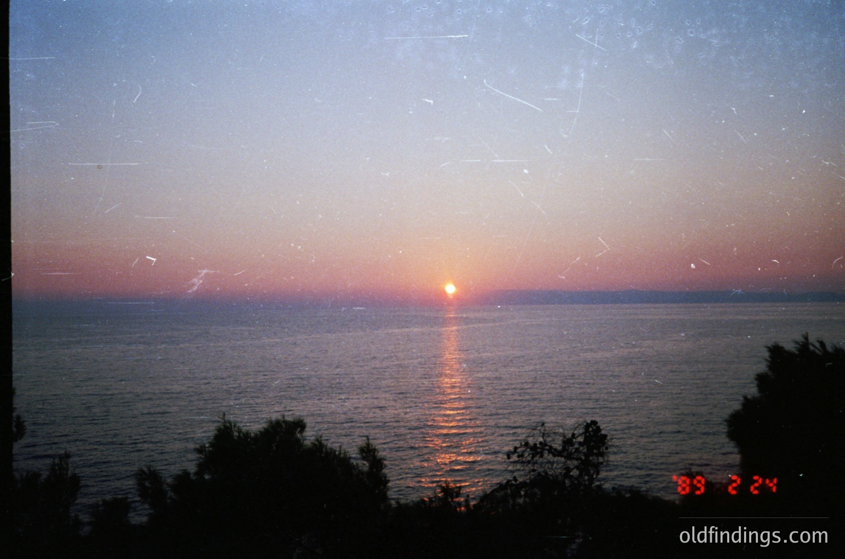 Vintage Polaroid of sunset over calm waters, dated 1989. Reflections on water and lens flare create a soft, nostalgic glow. Minimal horizon line with distant landmass.