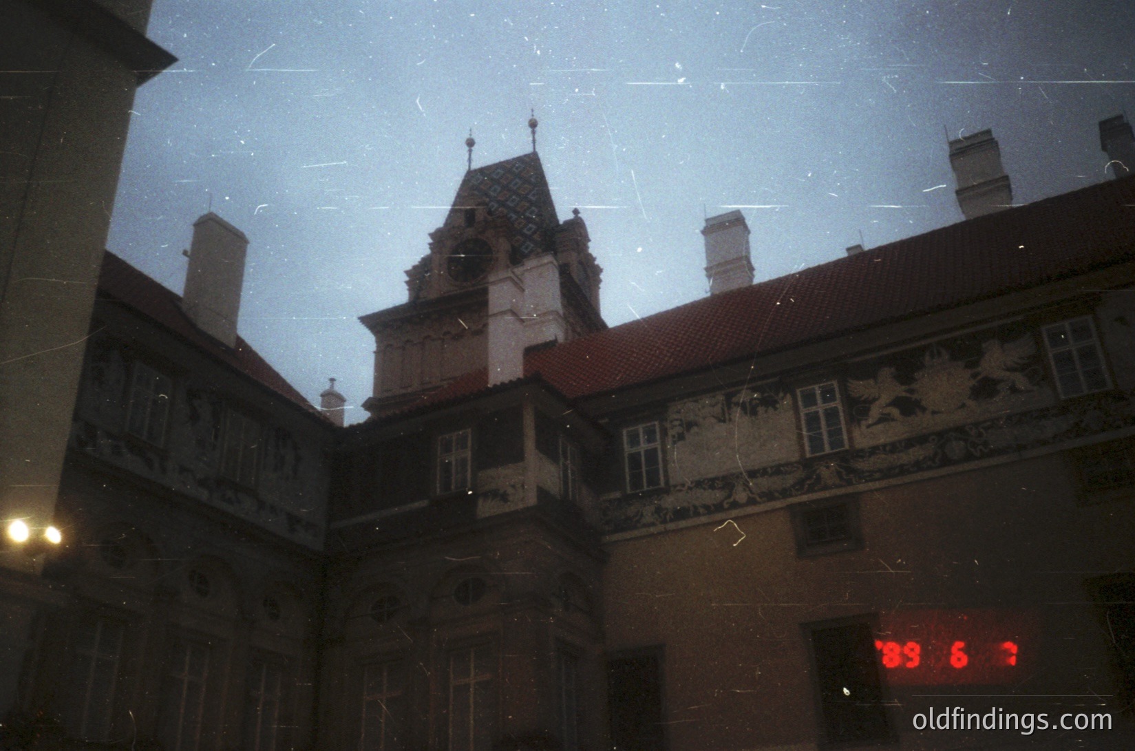 Vintage black-and-white photo of a European courtyard with ornate, multi-story building featuring Gothic Revival architecture—pointed arches, decorative brickwork, and a central tower with a steep, tiled roof. Visible street sign reads "39 67" in red. Likely 1950s–1970s urban setting.