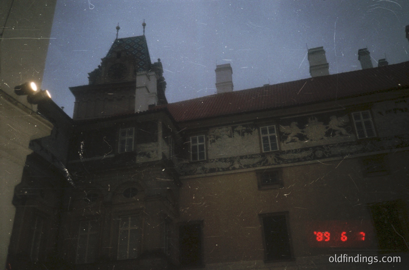 Reflection of a historic European building facade with ornate stone carvings—likely 19th-century—showing decorative motifs and steep gabled roofs. Overlaid timestamp "19:6:7" suggests a vintage or archival photo. Dark, moody lighting enhances architectural details.