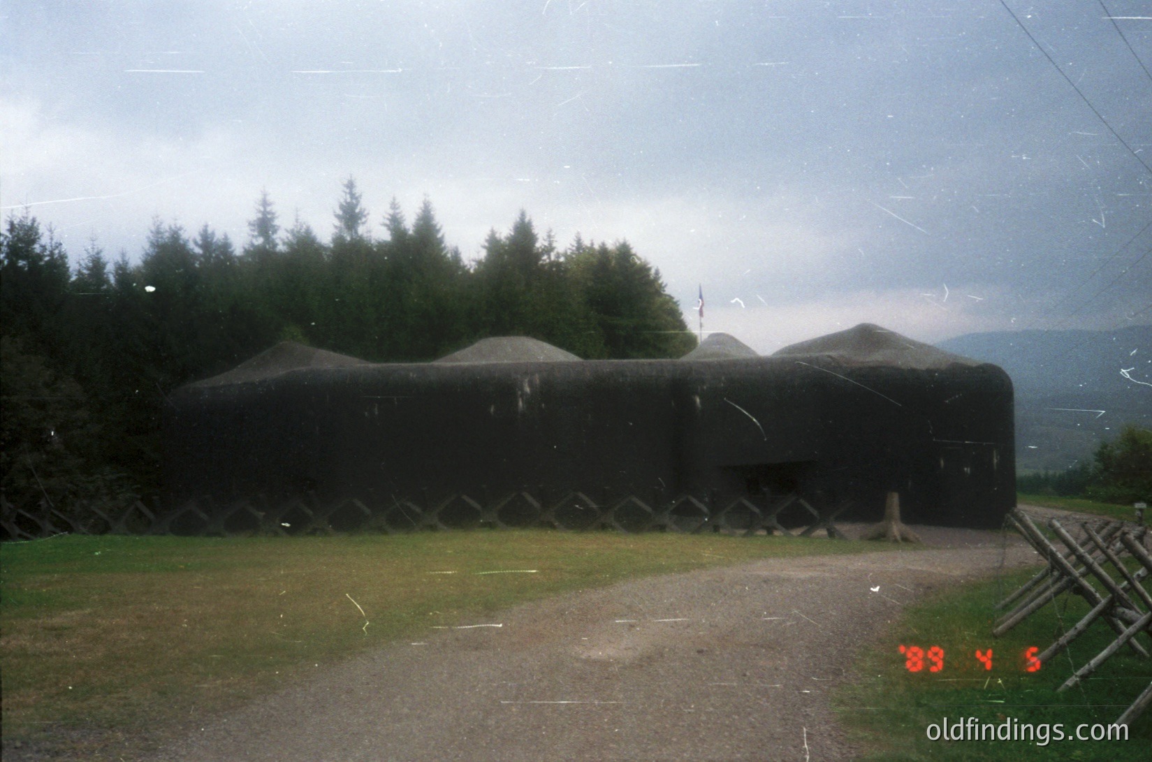 Concrete bunker with sloped roof and ventilation slits, set in a grassy field with forested background. Overcast sky suggests WWII-era Cold War defensive architecture. Timestamp "89 4 5" indicates possible 1989 date stamp.