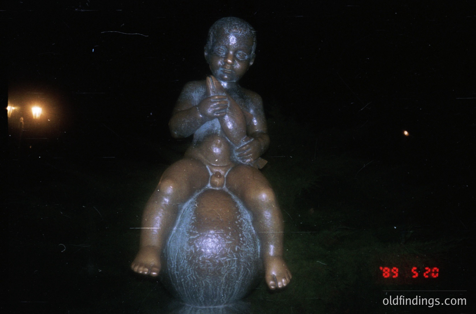 Bronze statue of a seated child playing a flute atop a spherical base, likely a public art piece. The low-angle lighting enhances texture and shadow. Stamp indicates vintage or archival quality, possibly from the late 20th century. Ideal for historical or artistic research.