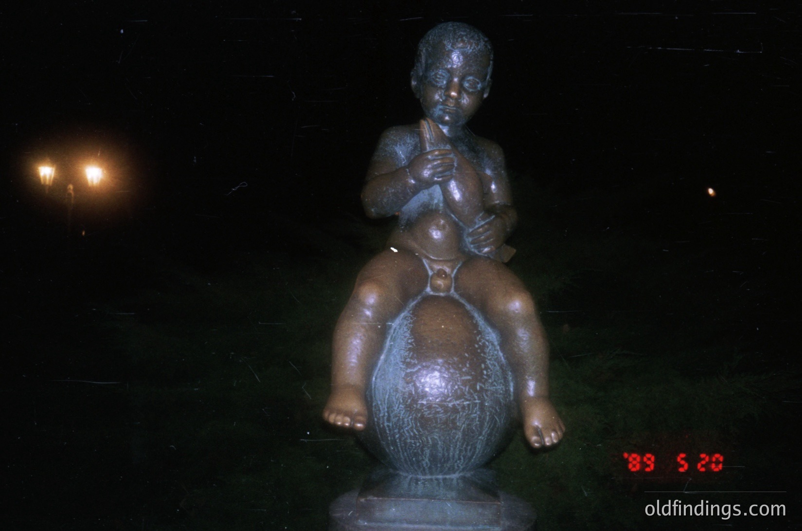 Bronze sculpture of a seated child playing a flute atop a spherical base, likely a globe. Low-angle nighttime lighting highlights texture and craftsmanship. Stamp indicates 1989, suggesting Soviet-era or Eastern Bloc artistic style.