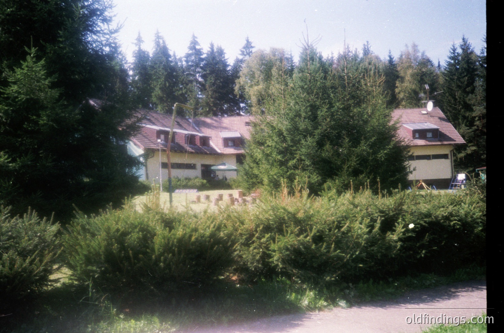 Two-story Soviet-era residential buildings with red-tiled roofs, set in a lush, forested area. Constructed from light-colored concrete blocks, featuring small windows and satellite dishes. Overgrown greenery and shrubs surround the perimeter. Likely Eastern Europe, mid-20th century.