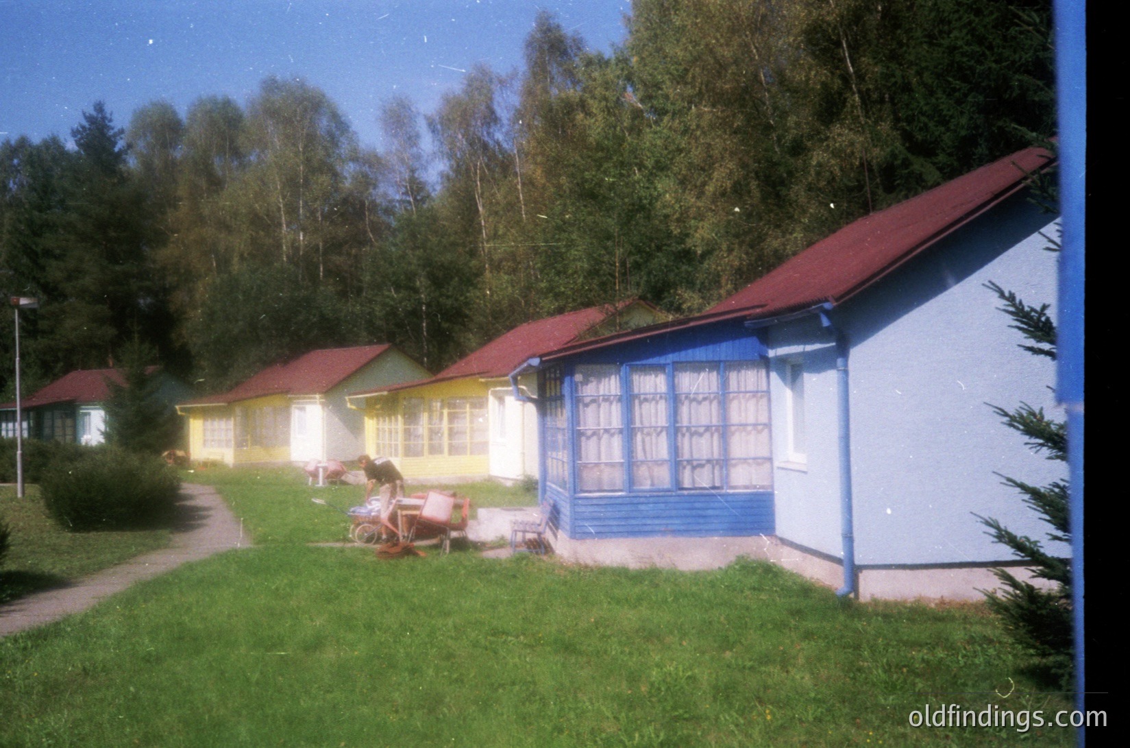 Vintage row of pastel-colored beach huts with red roofs, lined along a grassy path. Bright blue doors and windows contrast with yellow, white, and grey facades. Likely a seaside resort from the mid-20th century, possibly Eastern European.
