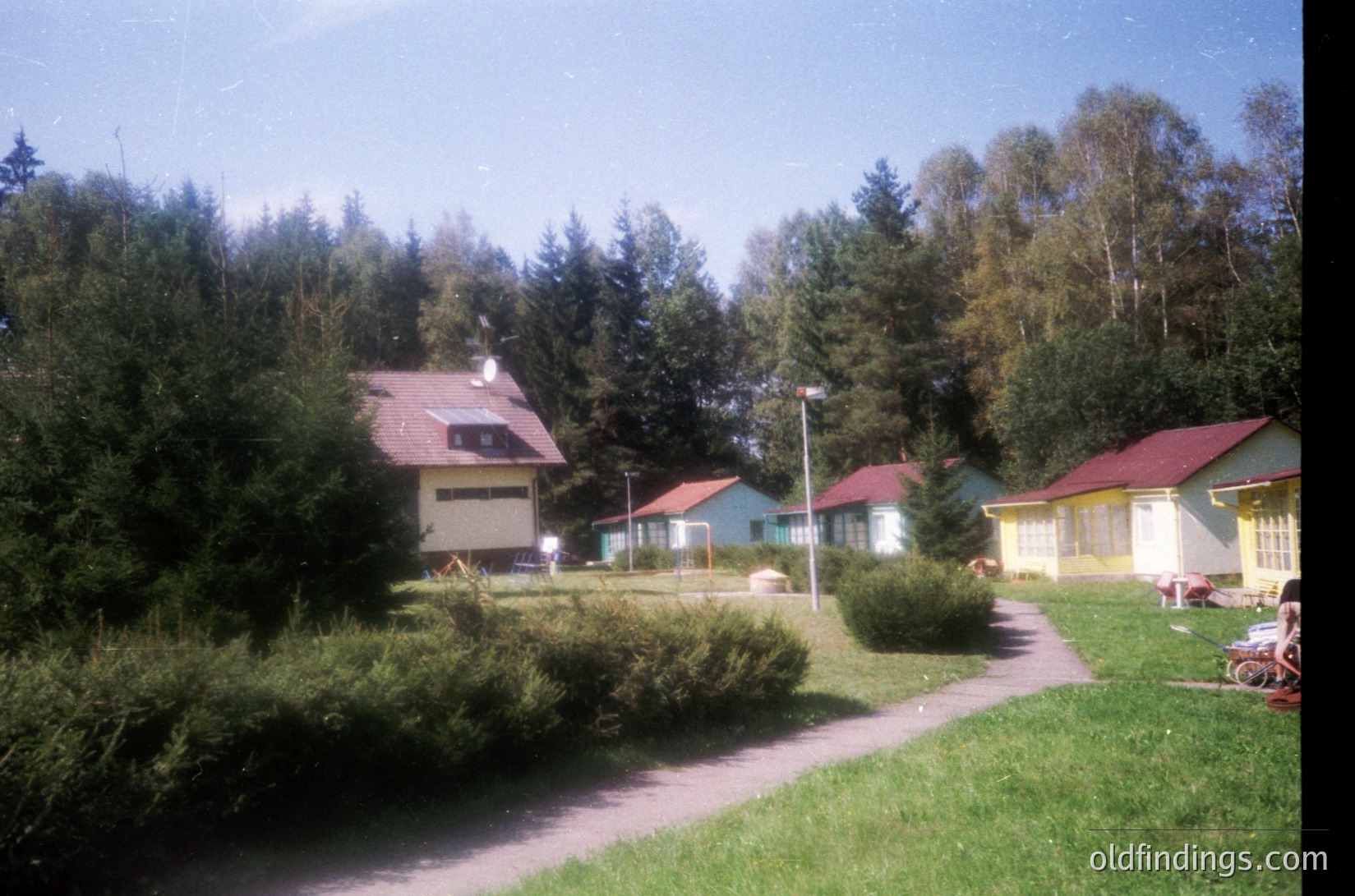 Vibrant 1970s-era resort complex nestled in a forested area, featuring pastel-colored bungalows with red roofs. Lush greenery, trimmed hedges, and a paved pathway dominate the scene. Classic Soviet-era architecture with simple, functional design.