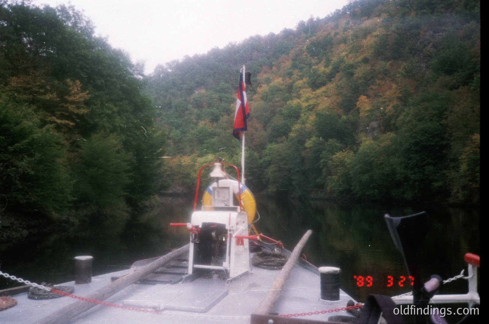 Vintage ferry crossing a narrow, forested river with autumn foliage. White vessel marked with red life rings and a red/white flag, bearing a digital speedometer reading 8.3 knots. Chain barriers and railings visible on deck. Likely Eastern European or Soviet-era design.