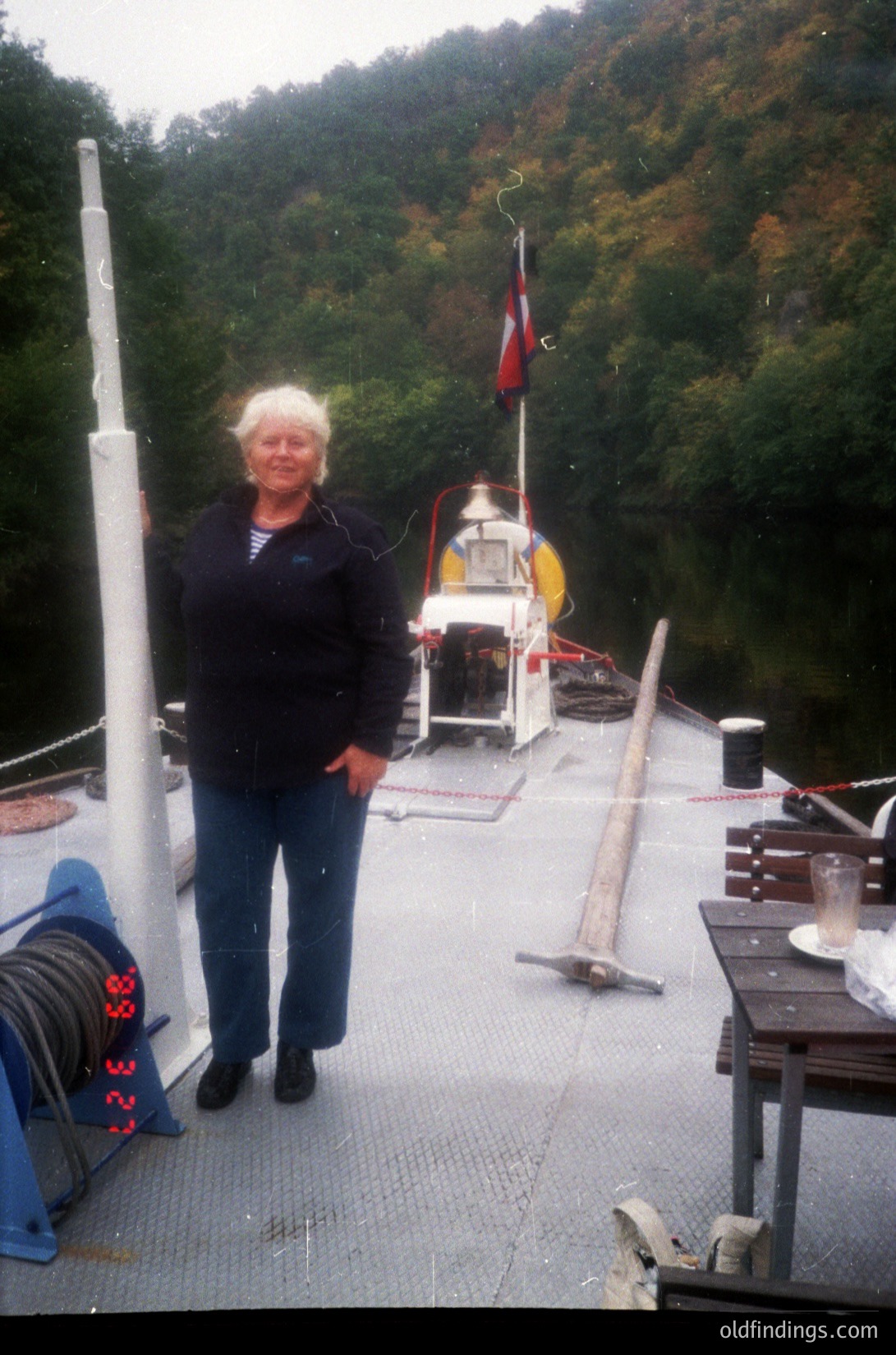 Woman in casual 1990s attire stands on a small wooden ferry deck, near a red-and-white flag. The vessel features a simple cabin and lifebuoys, docked near forested hills.