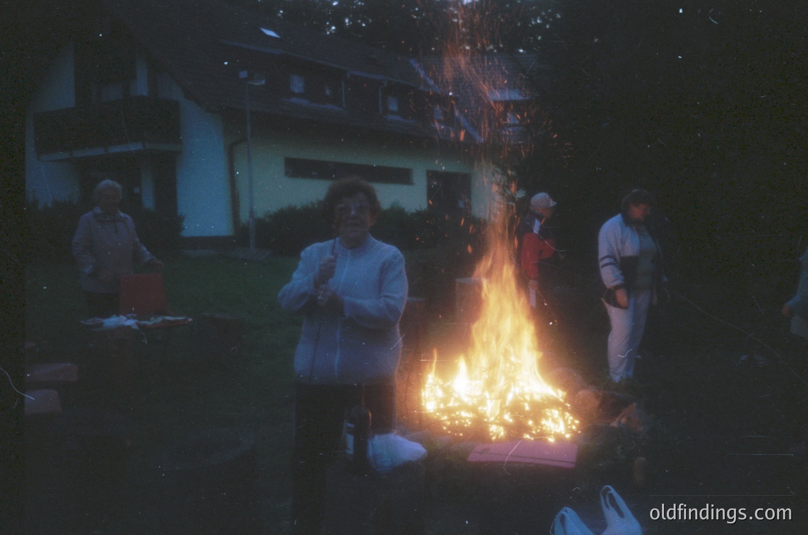 Vintage evening gathering around a bonfire in a residential backyard, likely 1970s–1980s. Group of adults in casual attire (tracksuits, sweaters) warming hands near flames. Suburban homes with brick/stone facades in background. Warm lighting contrasts with cool blue tones.