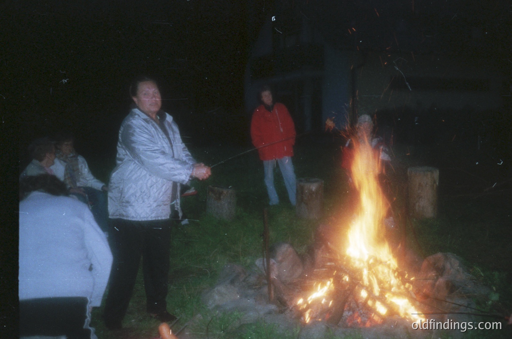 Nighttime outdoor gathering around a roaring bonfire. Four individuals in casual winter attire (puffer jackets, jeans) roast skewers over flames. Concrete structures and wooden barrels in background suggest a residential or park setting. Warm glow contrasts with dark surroundings, evoking communal warmth.
