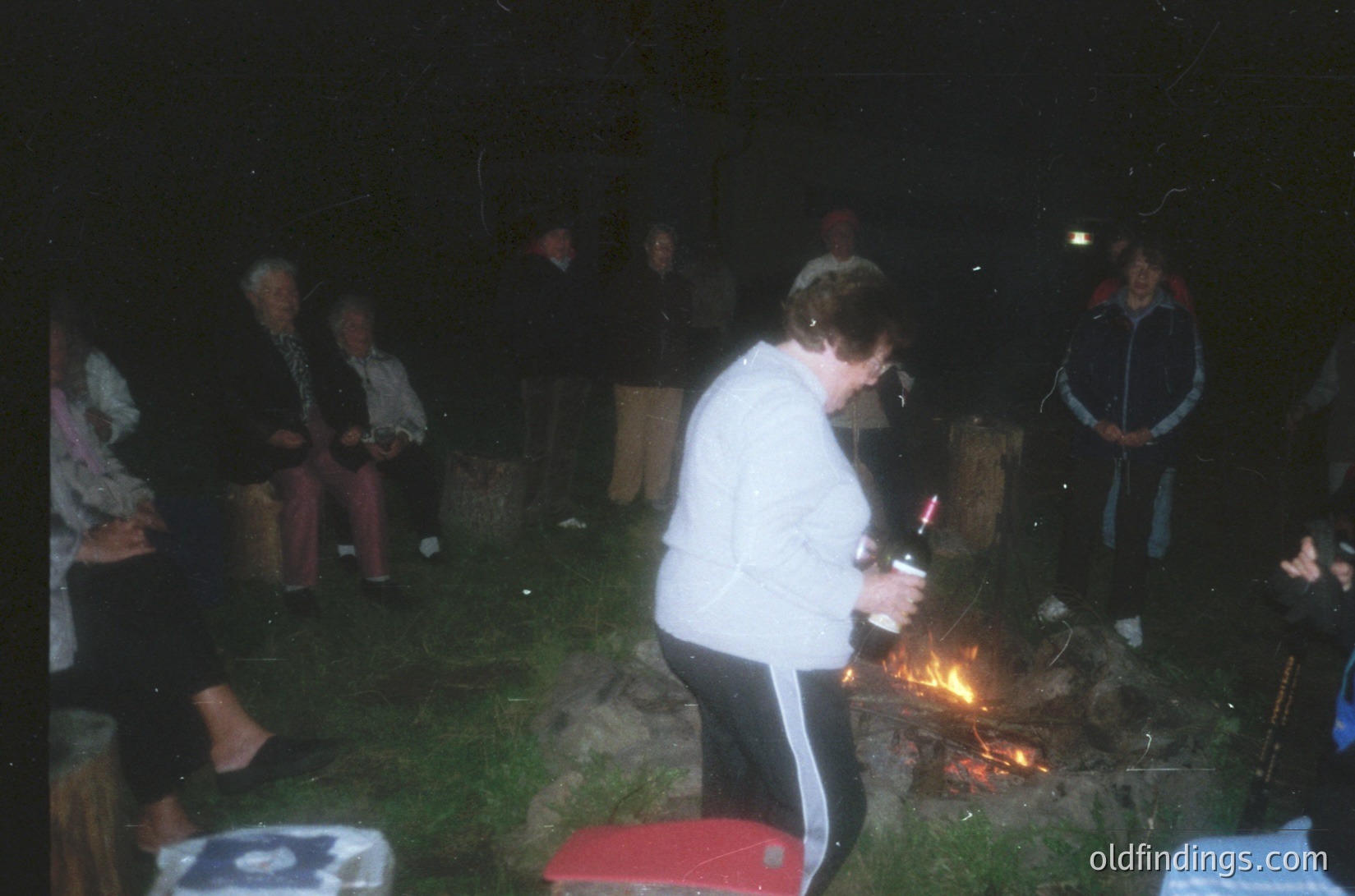 Nighttime outdoor gathering around a small bonfire. A person in a white jacket and black pants lights a candle or small firework near the flames. Others sit on stools or logs, engaged in conversation. Vintage camera quality suggests late 20th century. Casual, communal atmosphere.