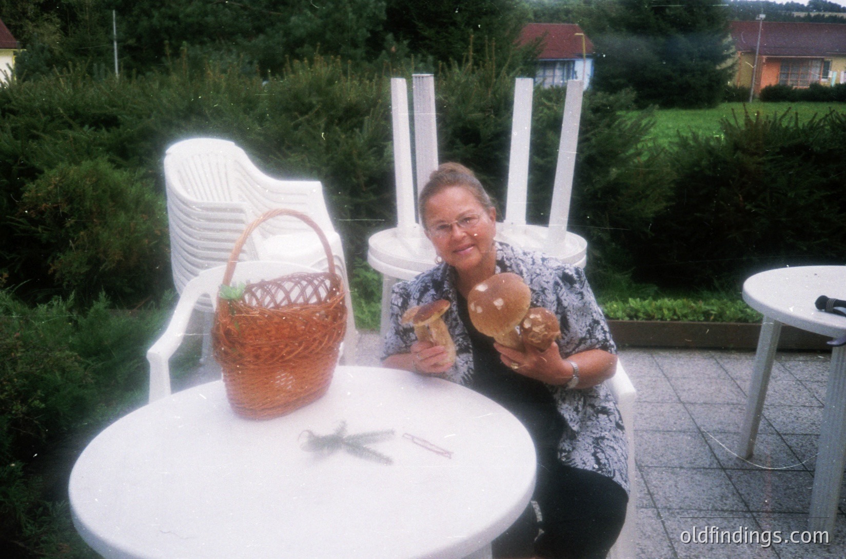Mid-20th century outdoor café scene featuring a woman in a floral blouse holding two pastries. White plastic chairs and tables, a woven basket on the table, and a blurred background of greenery and residential buildings. Likely