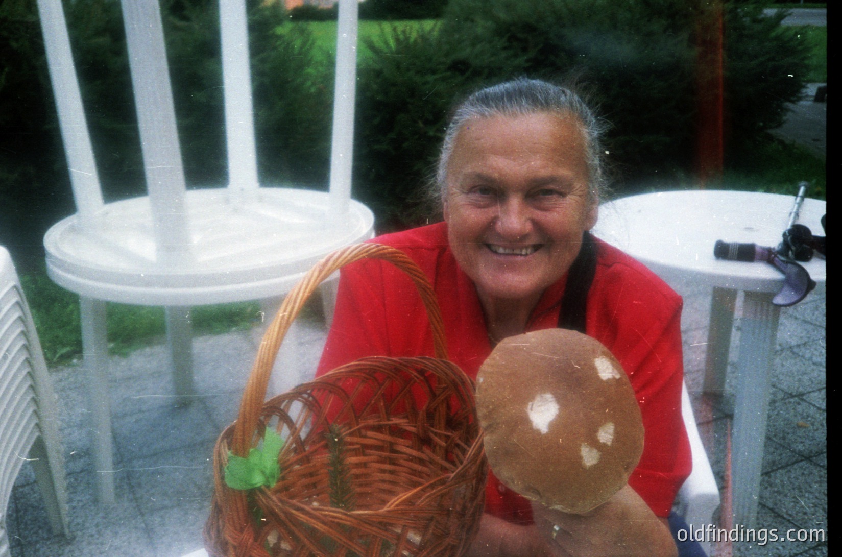 Elderly woman in a red sweater holds a large brown mushroom and woven basket, seated on a white plastic chair. Indoor/outdoor setting with glass doors and greenery in background. Likely 1990s–2000s, Western Europe/USA.