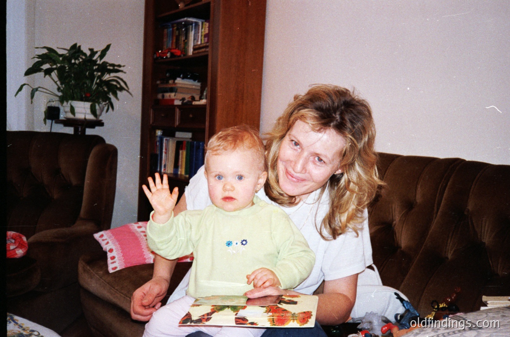 Vintage indoor portrait of a woman and toddler in a cozy living room, likely late 1990s–early 2000s. Woman in a white top holds a baby in a light green long-sleeve shirt, waving. Bookshelf with books, potted plant, and floral-patterned couch visible. Warm, nostalgic family moment.