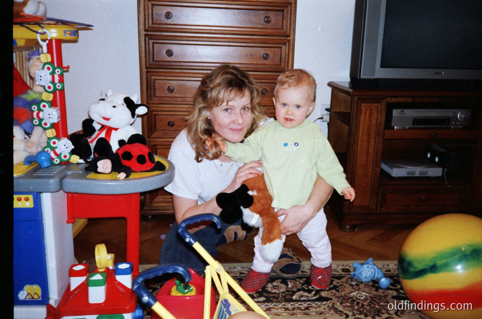 A woman and toddler pose indoors in a mid-20th-century home, likely 1980s–1990s. The woman holds the child in a light green sweater, while the child wears red shoes and a striped shirt. Surrounding them: a red-and-blue toy table with cow and ladybug toys, a wooden dresser, a TV, and a colorful exercise ball. Warm, lived-in family space.