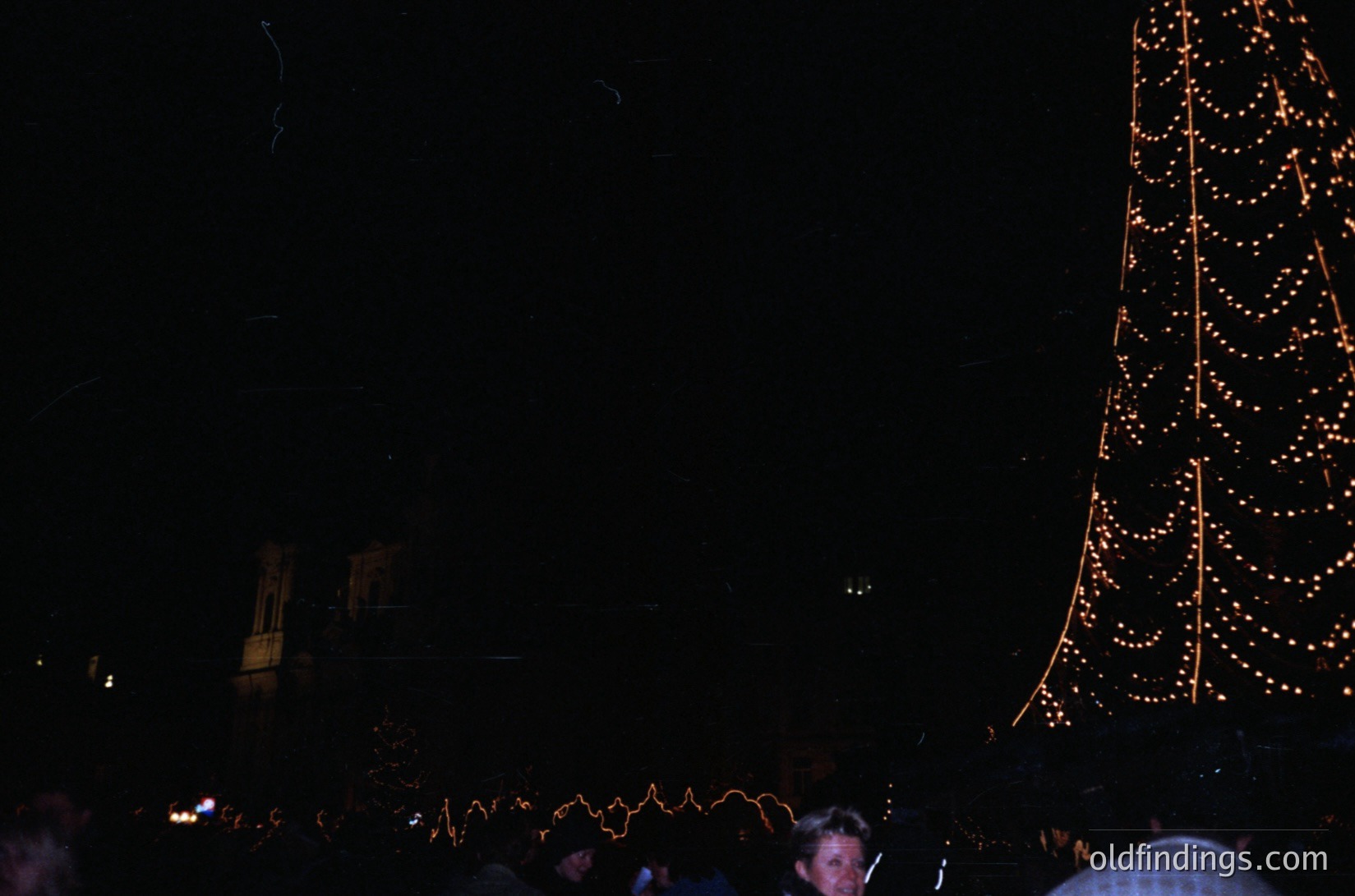 Vintage nighttime festive scene with illuminated tree and blurred crowd. Warm string lights outline a tall, conical structure, likely a Christmas tree. Distant buildings and silhouetted figures suggest an urban holiday event. Underexposed but captures communal atmosphere.
