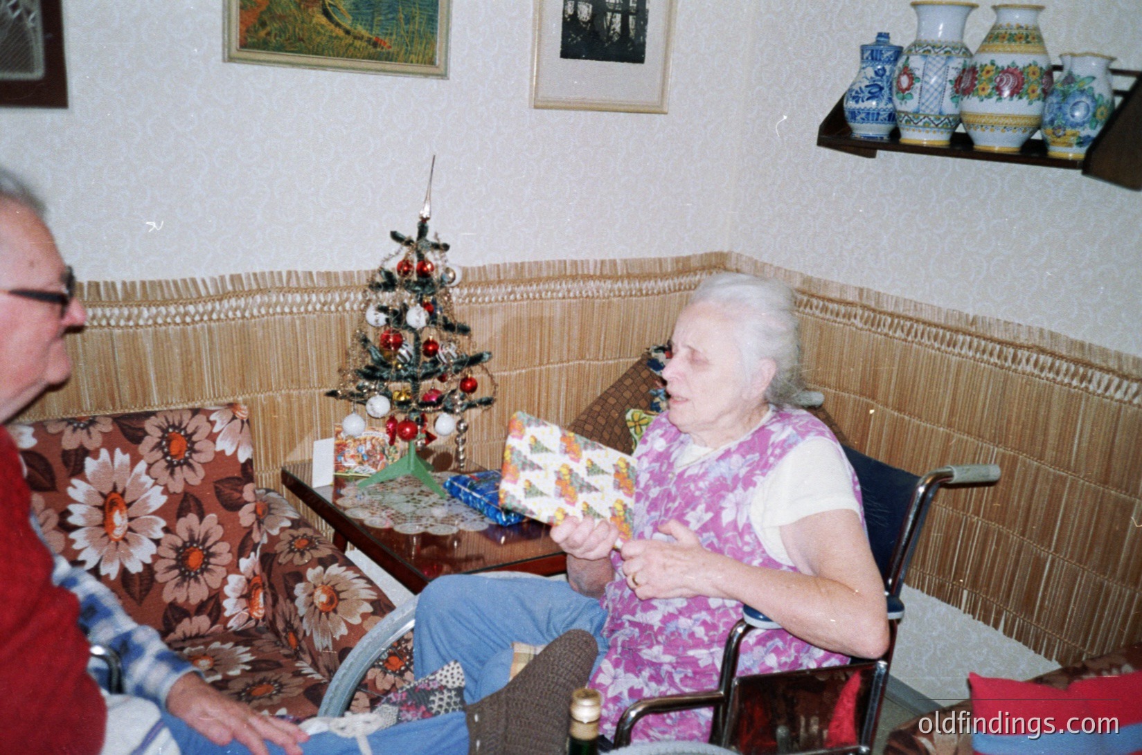 Vintage indoor scene featuring two elderly individuals in a cozy, textured room. A woman in a floral-patterned wheelchair holds a wrapped gift, while a man in a patterned sweater leans in. Decor includes a small Christmas tree, ceramic vases, and framed art. Warm, nostalgic 1970s–1980s home ambiance.