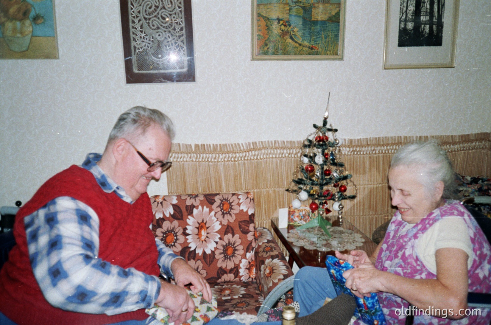 Two elderly individuals crafting in a cozy 1970s-era living room. The man, wearing a plaid sweater and glasses, and the woman, in a floral blouse, work on intricate paper decorations. Decor includes a Christmas tree adorned with glass ornaments and patterned wall hangings. Warm, earth-toned textiles and framed artwork enhance the nostalgic ambiance.