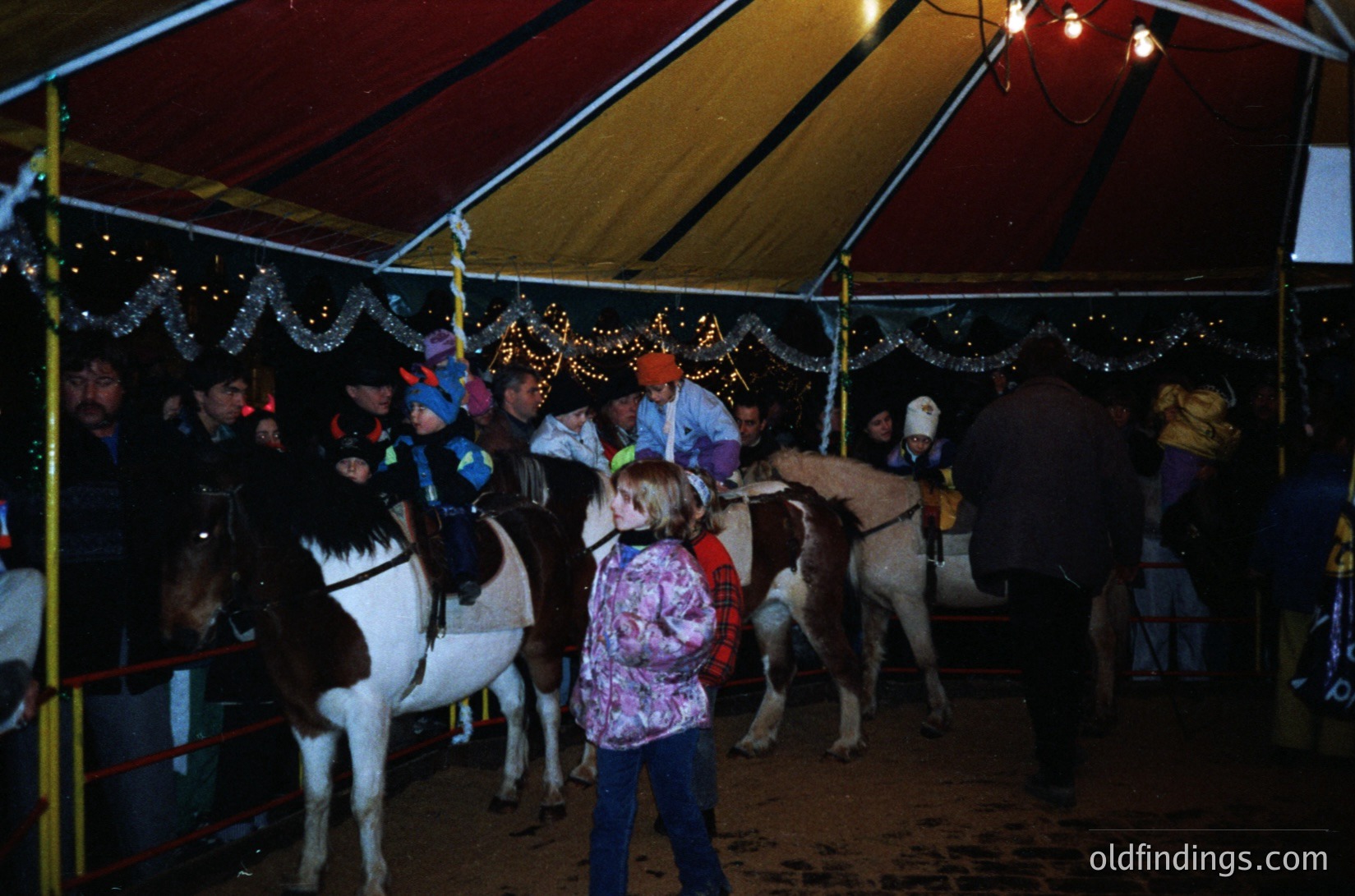 Vintage carousel tent with illuminated horses, likely from a 1970s-1980s fairground. Children and adults interact with painted carousel horses under festive string lights. Indoor/outdoor setting with striped tent canopy.