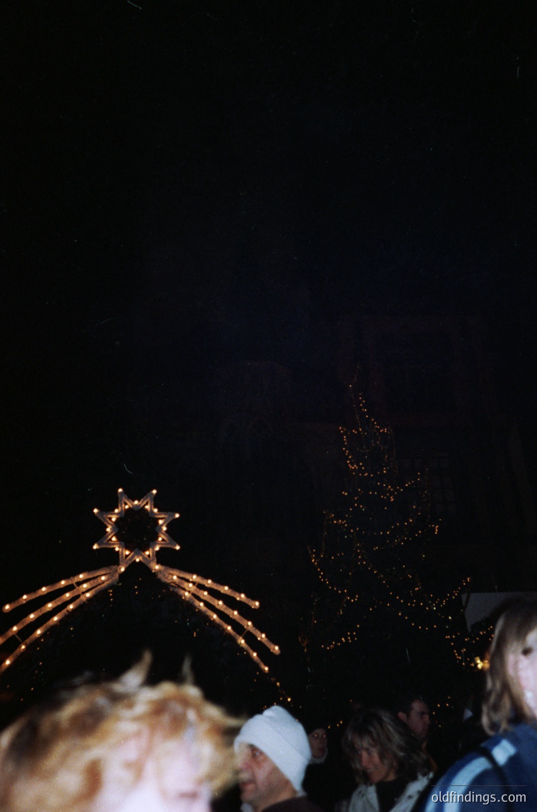 Blurred nighttime festive scene with illuminated tree and decorative arch. Warm golden lights contrast against dark background. Crowd silhouetted, suggesting holiday celebration. Likely 1990s–2000s era based on lighting style.