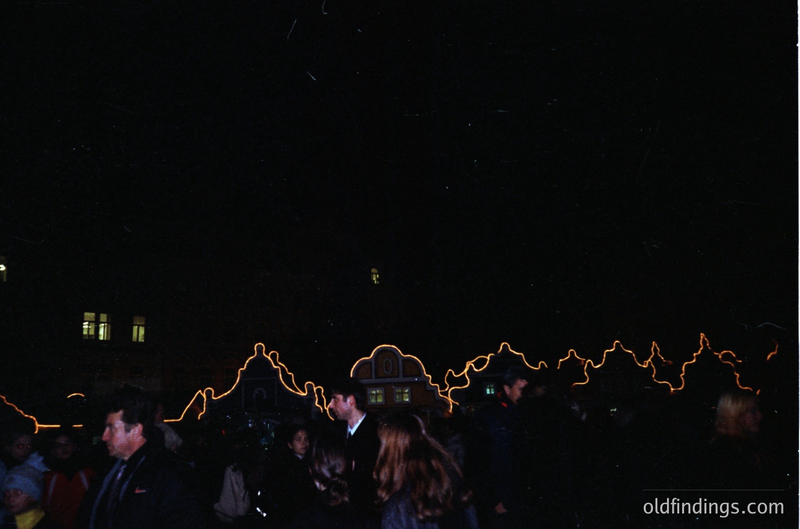 Vintage nighttime street scene with neon light trails from moving vehicles, capturing motion blur. Crowd of casually dressed people in mid-20th century attire, likely 1960s-1970s. Distinctive illuminated signage suggests urban setting, possibly a European city. Dynamic composition ideal for retro design or historical research.
