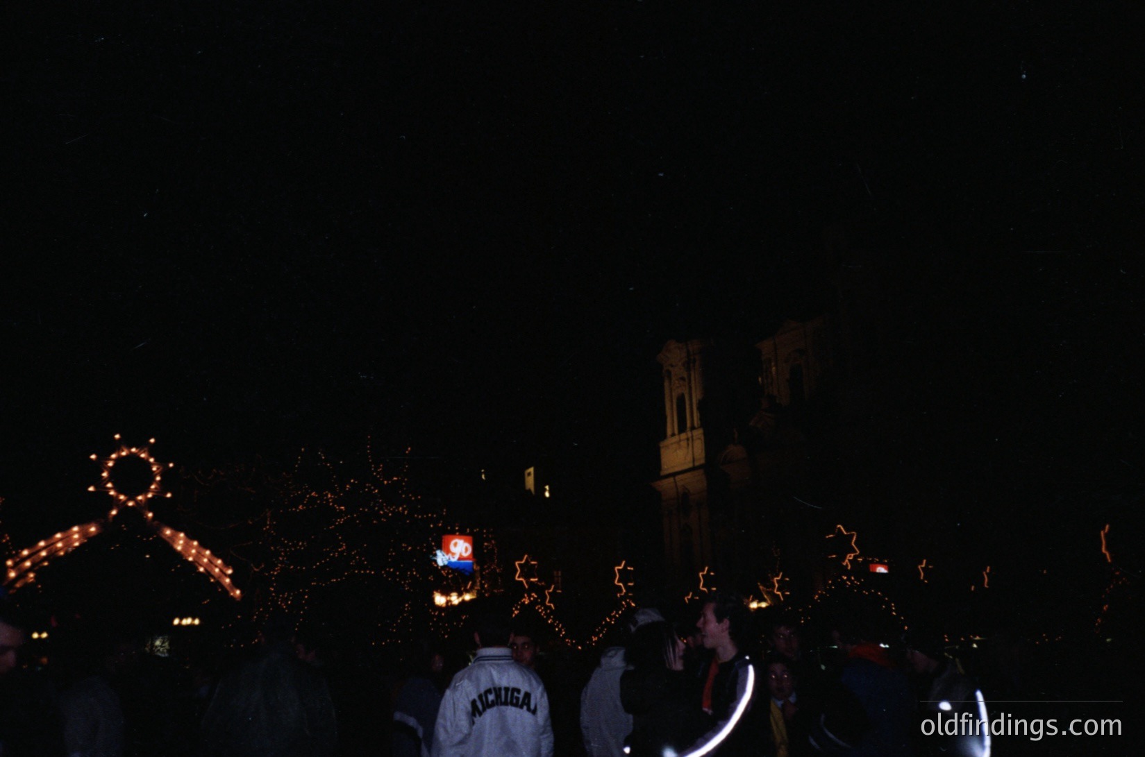 Nighttime crowd celebration with illuminated star decorations and a clock tower silhouette. Two individuals in Michigan-themed hoodies (front left) suggest a university event. Decorated trees and festive lights create a vibrant, communal atmosphere. Likely a 1990s–2000s college holiday party.