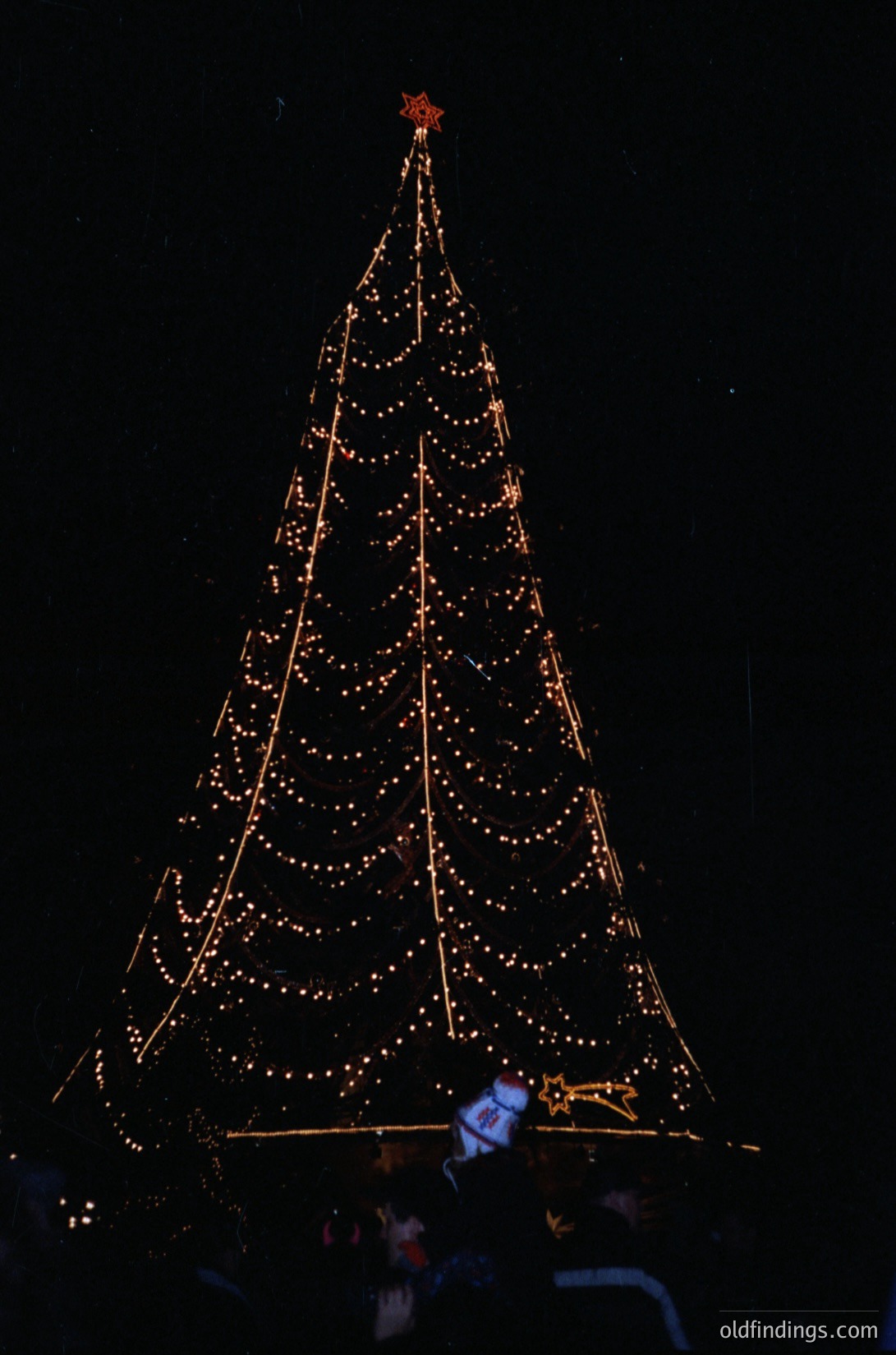 Impressive illuminated Christmas tree display with warm white and golden lights, crowned by a red star. Structure features horizontal beams forming geometric patterns. Crowd silhouetted below, suggesting festive event atmosphere. Likely modern holiday lighting design for urban or public space.