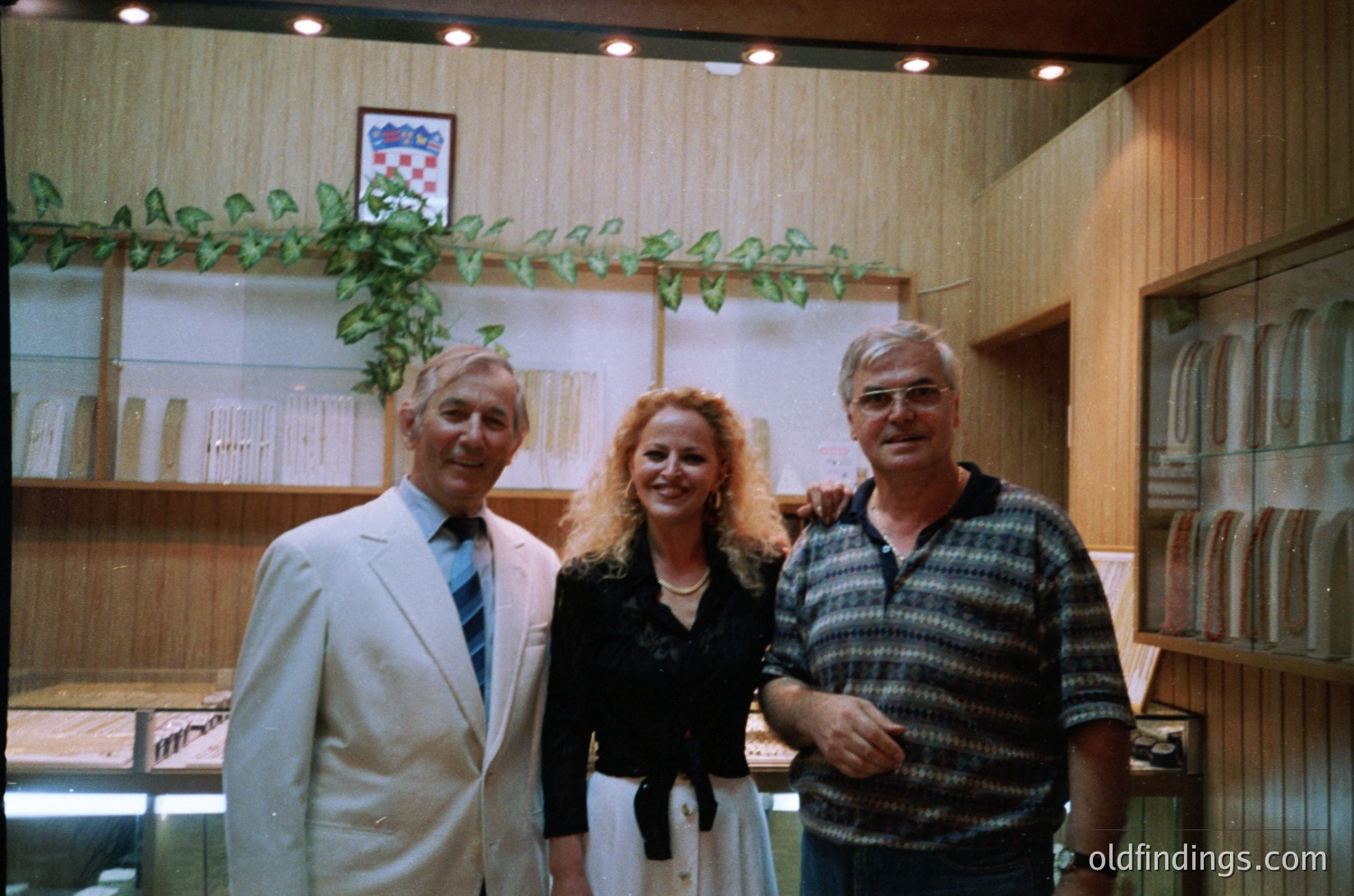 Three individuals pose indoors in a formal setting, likely a 1970s–1980s exhibition or gallery. The man on the left wears a light-colored suit with a striped tie, the woman a voluminous blouse with a draped skirt, and the man on the right a patterned sweater. Wood-paneled walls and framed displays suggest a cultural or academic venue. Warm lighting and vintage color tones enhance nostalgic appeal.