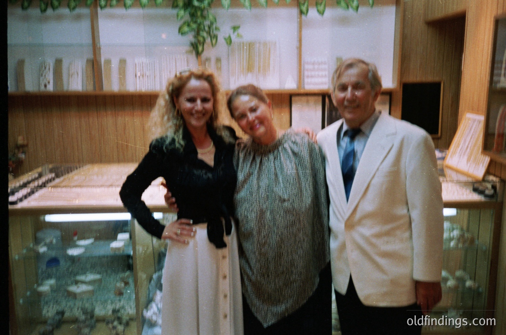 Three individuals pose indoors in a well-lit, mid-century setting. The woman on the left wears a sleeveless black top with a white skirt, the center figure a patterned blouse with a shawl, and the man on the right a white blazer with a striped tie. Behind them, a large aquarium with fish and a display case with model ships suggest a maritime or nautical theme. The interior features wooden paneling and framed artwork. [Vintage family portrait in maritime-themed interior ]