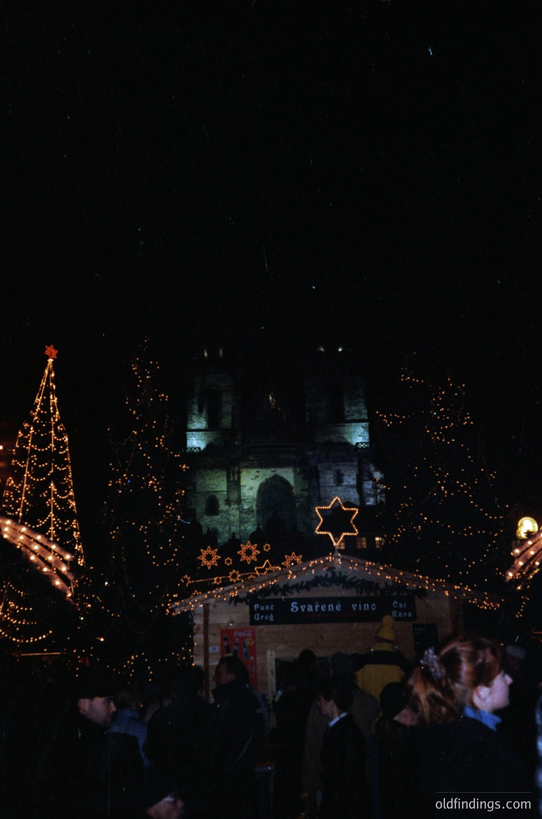 Nighttime festive scene featuring illuminated Christmas decorations. A large, ornate tree adorned with warm lights and star ornaments dominates the left. In the background, a historic building with arched windows and a clock tower is lit from within, casting a soft glow. Decorative star and gear-shaped lights frame a sign reading *"Piv Svarene vinn"* (likely a beer festival). Crowds gather below, suggesting a lively holiday market atmosphere.