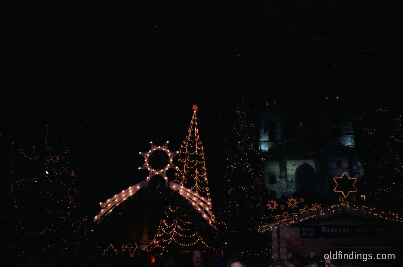 Vintage-style nighttime holiday display featuring illuminated Christmas tree, star, and menorah-inspired lights. Architectural details suggest a European castle-like setting. Likely 1970s–1990s holiday event.